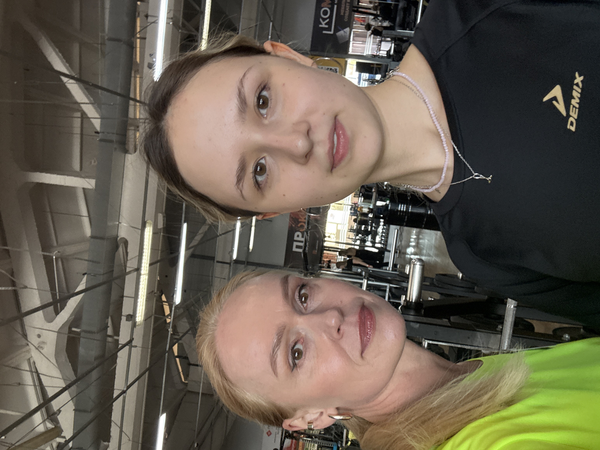 Two women pose for a selfie indoors. The younger woman is on top, wearing a black DEMIX t-shirt and a beaded necklace. The older woman, with blonde hair, is on the bottom wearing a bright green top. The background shows gym equipment in what appears to be a gym environment with exposed ceiling beams and industrial lighting. "KOM" is visible on a sign in the background, and a banner with Cyrillic script is also present. The location is likely Chaikovsky, Russia.