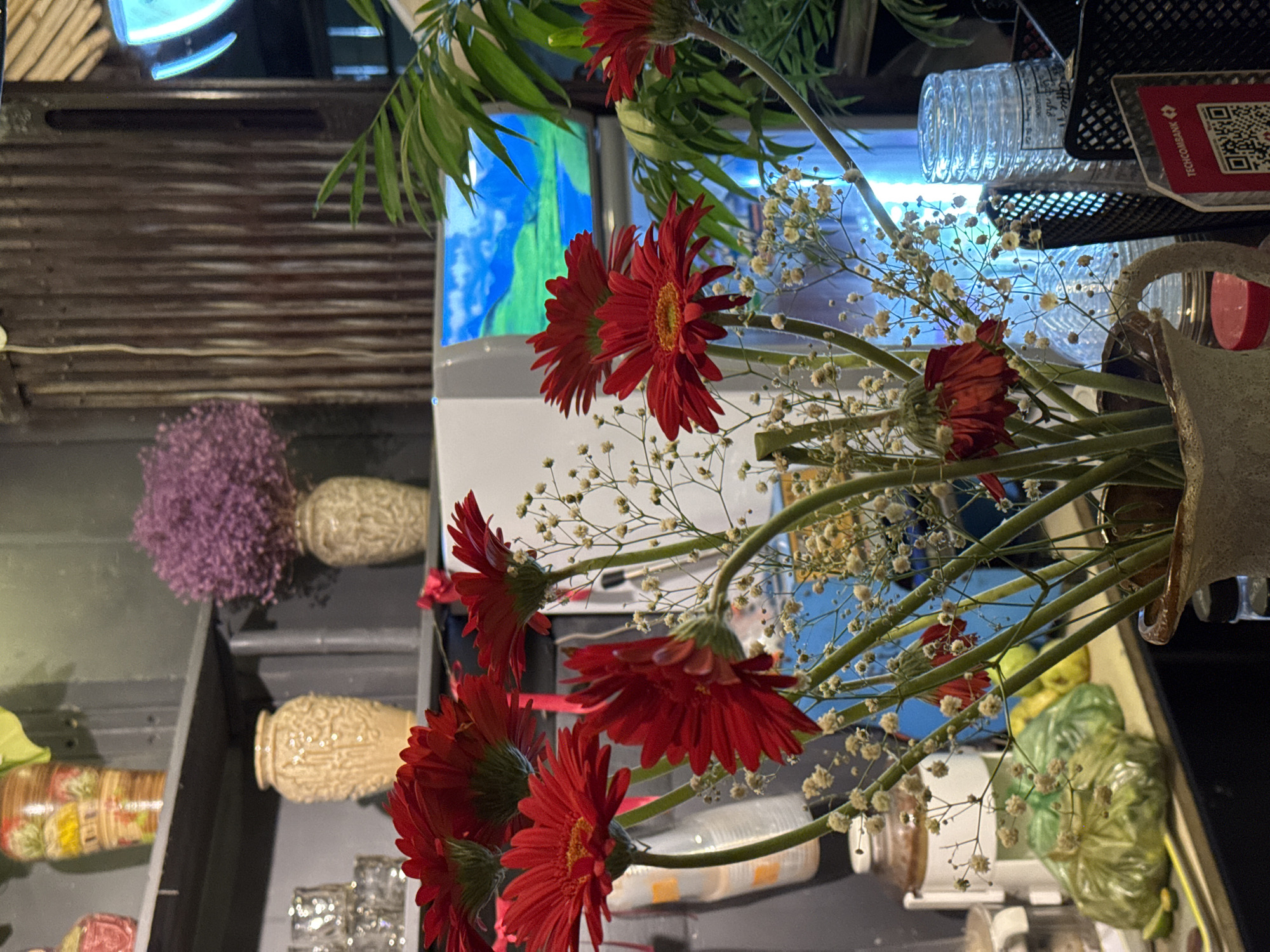 A still life photograph captures a vase of vibrant red gerbera daisies with delicate baby's breath flowers. The bouquet is arranged in a textured, beige ceramic vase. In the background, a television displays a landscape scene. To the left of the television, a corrugated metal wall is visible. Further back on a shelf, a cluster of purple flowers in a beige vase and another beige vase are present. The scene is likely set indoors, possibly in a cafe or home, given the arrangement of objects and decor. The photograph does not provide any specific indicators to pinpoint a location in Da Lat, Vietnam.
