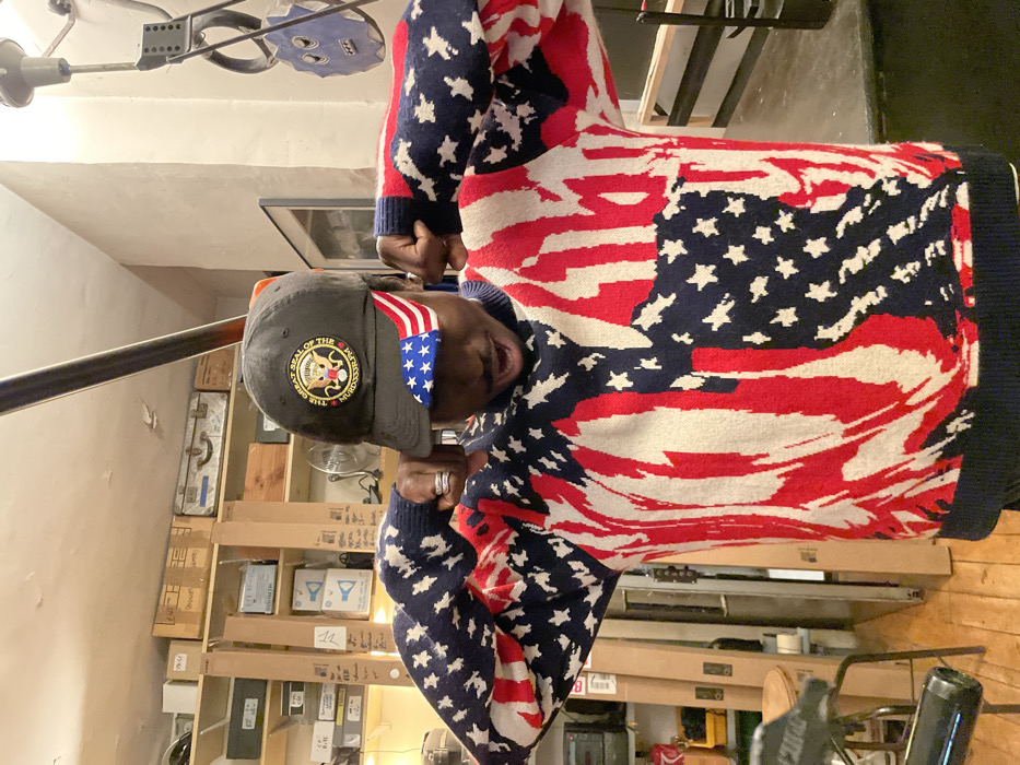 An individual, presumably a man, wearing a sweater patterned with an American flag design, a black baseball cap adorned with an emblem, and a flag headband. He is indoors and slightly tilted to the left in the shot. Behind him is a cluttered wall with stacked boxes and storage items. A metal rail runs diagonally across the upper left. The picture is rotated counterclockwise.
