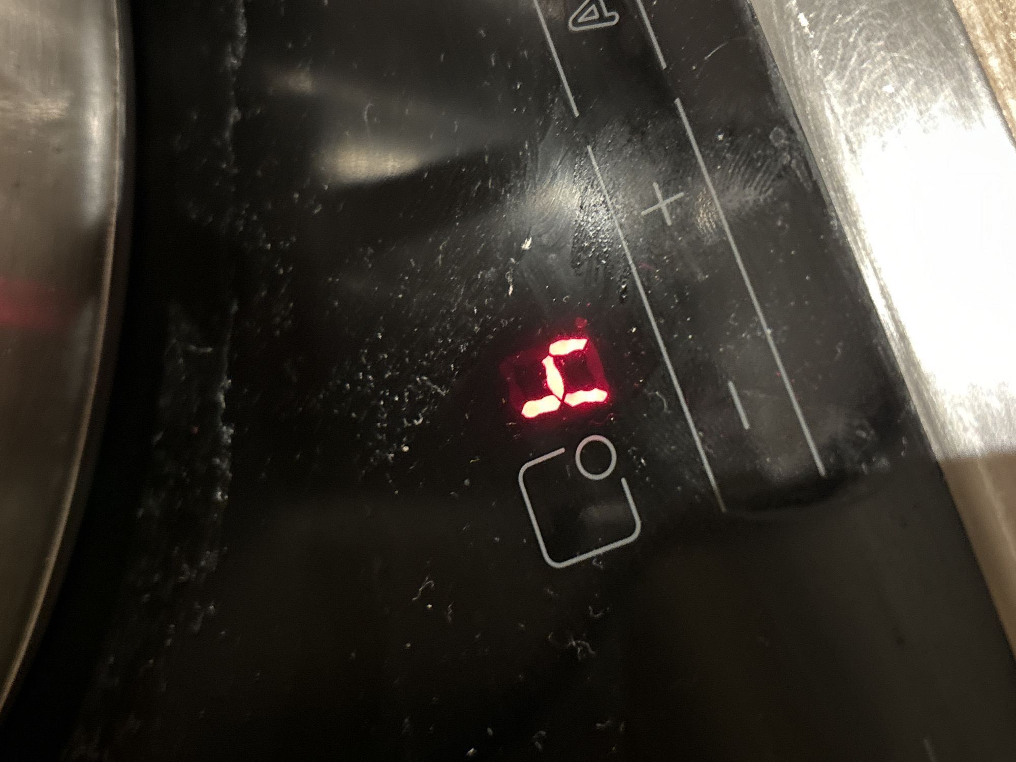 The image shows a close-up of a black stovetop with a red digital display showing the number "5". A white outline of a cooking pot symbol is located below the number. Control markings, including a "+" sign and the number "4", are visible at the top right of the image. A portion of a pot is visible on the left side of the image. The stovetop surface is dirty with what appears to be food debris. There is no information in the image to determine a location in Budva, Montenegro.
