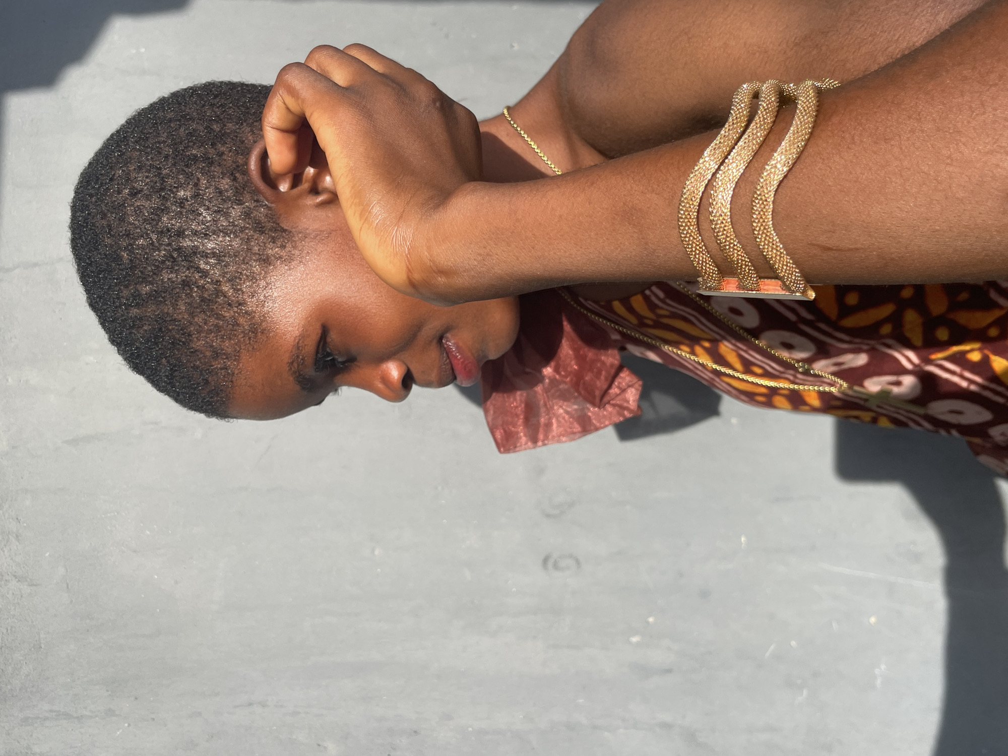 A medium shot, taken outdoors in Badagry, Nigeria, captures a person of color, likely a woman, in profile. The subject has short, dark, closely-cropped hair and is looking downwards with a gentle, serene expression. Their face and arms are illuminated by direct sunlight, highlighting their skin and giving it a luminous quality.
One hand is raised, with fingers gently touching the side of their head near the ear. The other arm is bent, showcasing a distinctive gold bracelet composed of three wavy, textured bands. A thin gold chain necklace is visible around their neck. The person is wearing a garment with a patterned fabric in tones of brown, yellow, and white, featuring a solid reddish-brown collar or detail.
The background consists of a plain, light gray or light blue-gray surface, possibly a paved ground or wall, with subtle texture. Strong, distinct shadows are cast by the subject and another unseen object on the ground, indicating bright, clear daytime conditions. There is no visible text in the image.