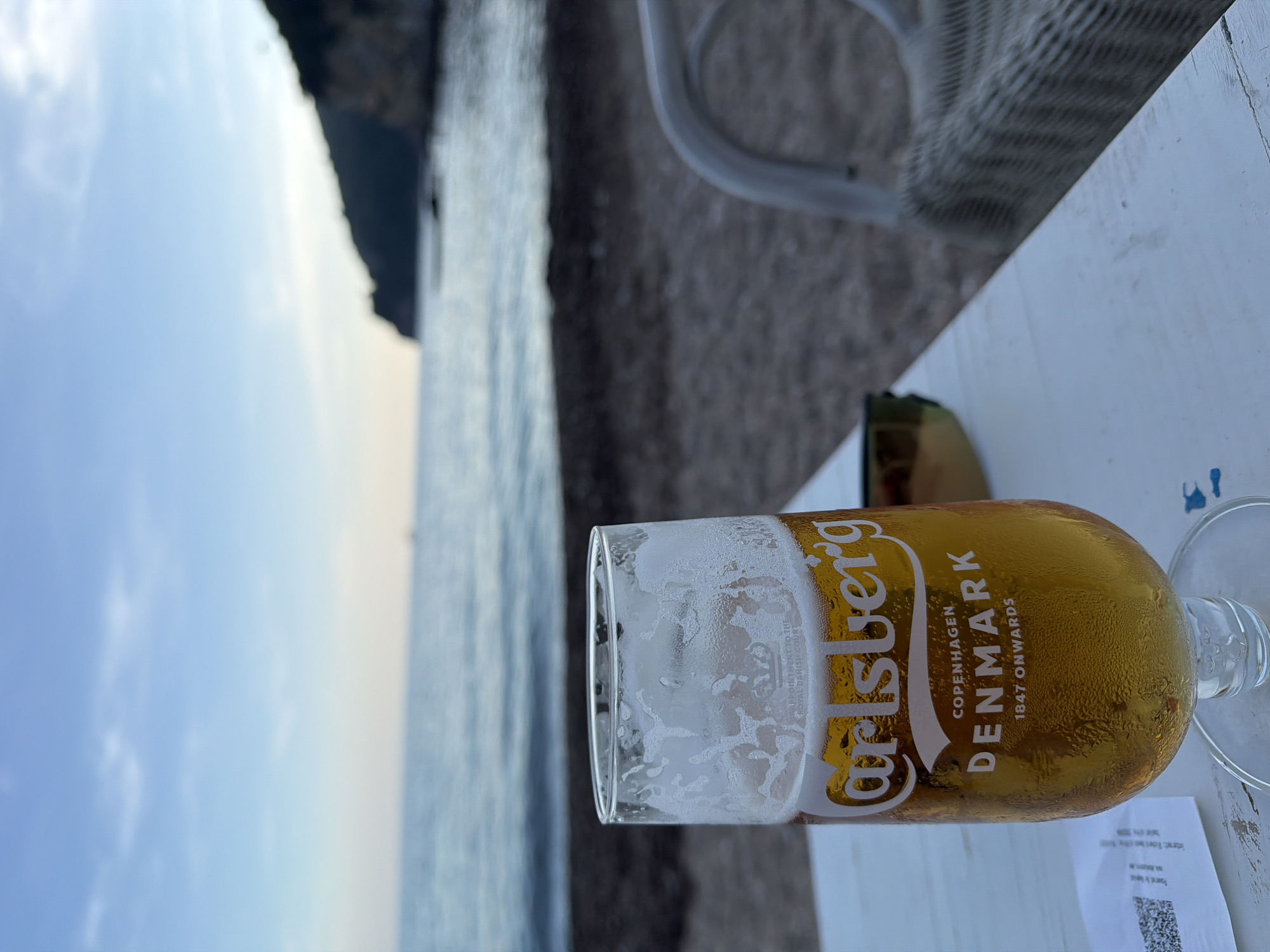 The original image, if correctly oriented, shows a glass of beer on a table set against a coastal backdrop, likely in Budva, Montenegro.
The main subject is a tall glass of Carlsberg beer, approximately half full, positioned on a light-colored, possibly wooden, table. Condensation covers the outside of the glass, and foam rings cling to the inside, suggesting the beverage is cold. To the left of the glass, a portion of dark-lensed sunglasses rests on the table. To the right of the glass's base, a crumpled white paper or receipt is visible, featuring a clear QR code and some unreadable text.
The visible text on the glass includes the prominent "Carlsberg" logo, followed by "COPENHAGEN," "DENMARK," and "1847 ONWARDS." Partially visible text below the logo also reads "APPOINTMENT TO THE ROYAL DANISH COURT."
In the mid-ground, a blurred, light-colored wicker or rattan chair is situated to the right of the table. The ground surface beyond the table and chair appears dark and textured, possibly pebbles or sand. The background features a serene body of water, likely the sea, which is a muted blue-grey and appears calm. In the distant left, a dark, rugged landmass or cliff rises from the water, partially obscured by atmospheric haze. The sky above is a soft, pale blue with a few faint clouds, indicating late afternoon or early evening.
The scene suggests a relaxed setting at an outdoor establishment by the sea, offering a tranquil and refreshing moment.