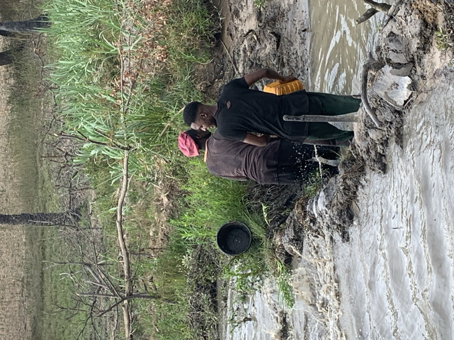 Two individuals stand along a muddy riverbank, with a black bucket positioned near the water's edge. The person in the foreground wears a pink head covering and a brown shirt. The other person is dressed in a black shirt and holds a yellow container. The river flows rapidly, causing splashes against the muddy bank, with vegetation growing on the opposite side. A thin tree with sparse branches is visible on the left of the frame.