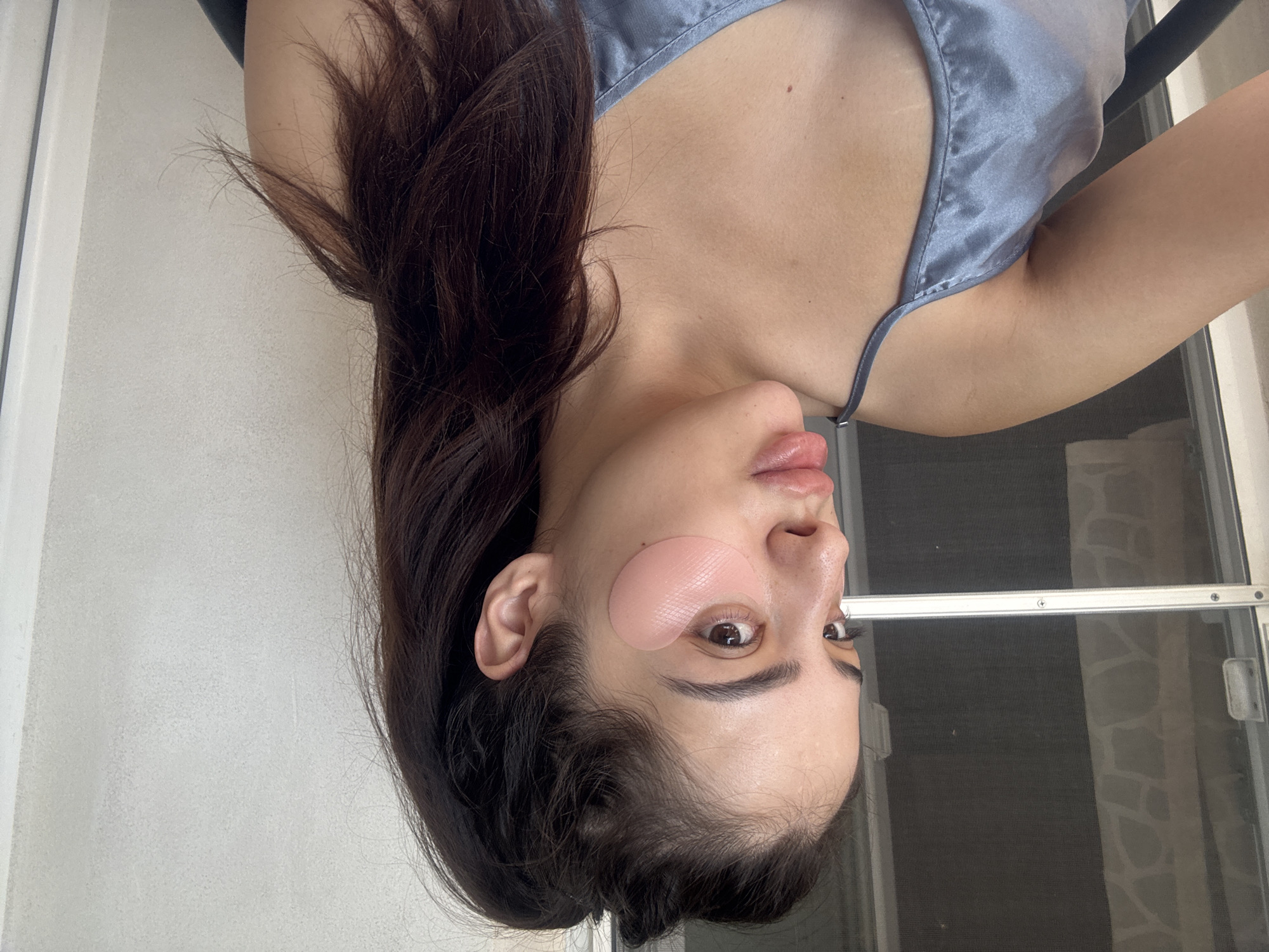 The image displays a close-up portrait of a person, likely a woman, with long, dark brown hair and light to medium skin tone. The photograph is rotated 90 degrees counter-clockwise. She is wearing a light grey or blue satin camisole with thin straps.
Her face is the central subject, featuring prominent eyebrows and full, slightly glossy lips. A pink, textured under-eye patch is applied beneath her right eye (viewer's left), partially obscuring it. Her visible left eye is open and looking towards the right side of the frame. The activity taking place appears to be part of a skincare routine, specifically the application of eye masks.
The background on the left side of the frame is a plain, light-colored wall, while the right side reveals a window or screened door. This window or door has a dark mesh screen and a horizontal silver-colored frame. The lighting suggests it is daytime. The setting appears to be an indoor or balcony environment, possibly in a home in Plovdiv, Bulgaria. No discernible text is visible in the image.