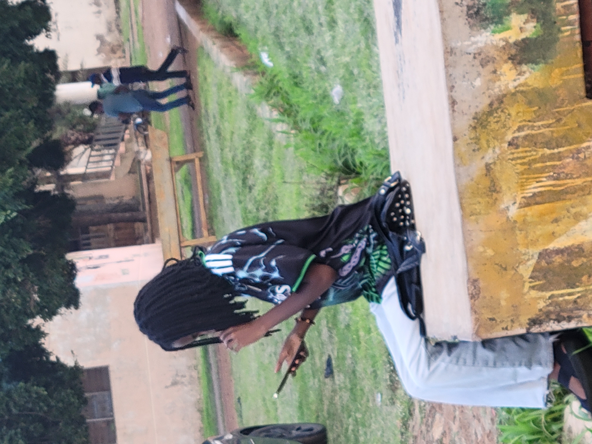 In the city of Jos, Nigeria, a young person with dark, braided hair sits on a low concrete structure. They are wearing a black t-shirt with a graphic design featuring white and green accents, light blue jeans, and black sandals. The person is holding a smartphone in their left hand and appears to be interacting with it. A black bag with stud detailing is placed on the concrete next to them. In the background, two men are walking away from the camera. One is wearing a blue t-shirt and jeans, and the other is in a dark uniform with a blue cap. They are on a path surrounded by green grass and trees, with a building visible in the distance. The overall environment suggests an outdoor public space.