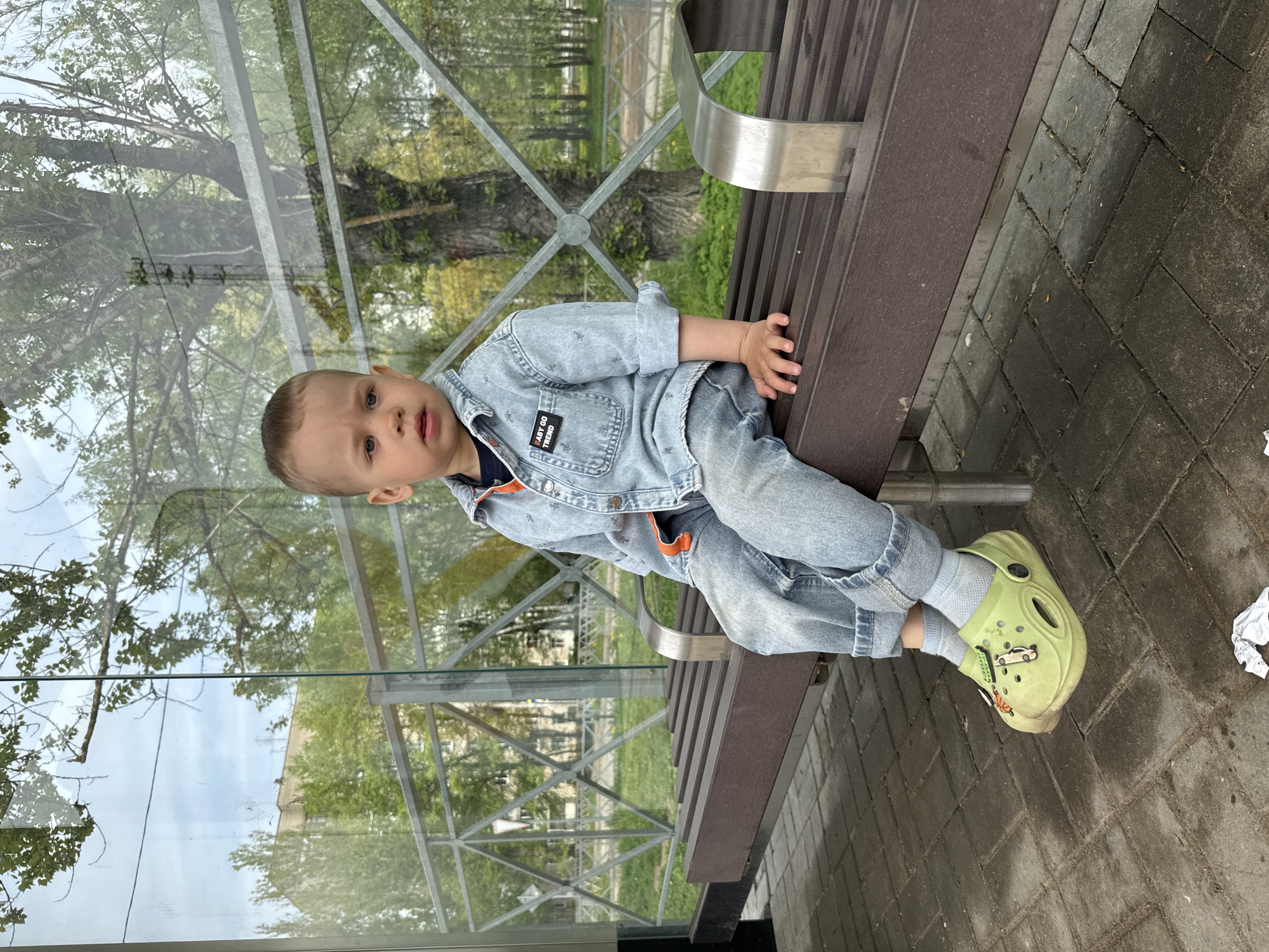 A young child is sitting on a brown bench in what appears to be an urban area. The child is wearing a denim jacket and jeans, paired with green Crocs-style shoes. The bench is situated near a glass structure that reflects trees and buildings, suggesting an outdoor setting. The ground is paved with interlocking bricks. The background shows foliage and architectural elements, indicative of the Rusanovo district in Russia.