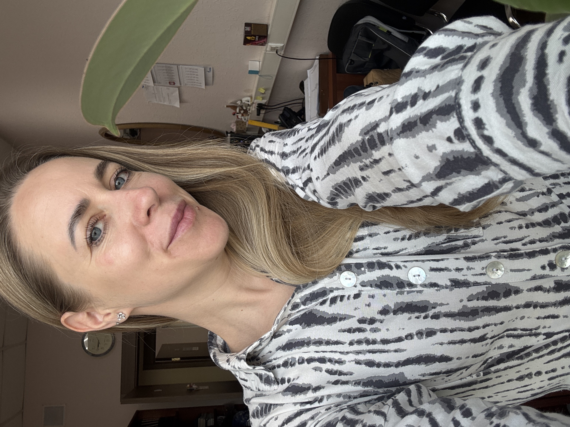 A woman with blonde hair and blue eyes poses for a selfie indoors. She wears a gray and white patterned blouse with visible buttons. Behind her, a partially visible clock hangs on the wall. The environment also includes a desk with various items on it and a black office chair. A green leaf is present in the foreground. The image does not explicitly state that the location is in Chaikovsky, Russia.