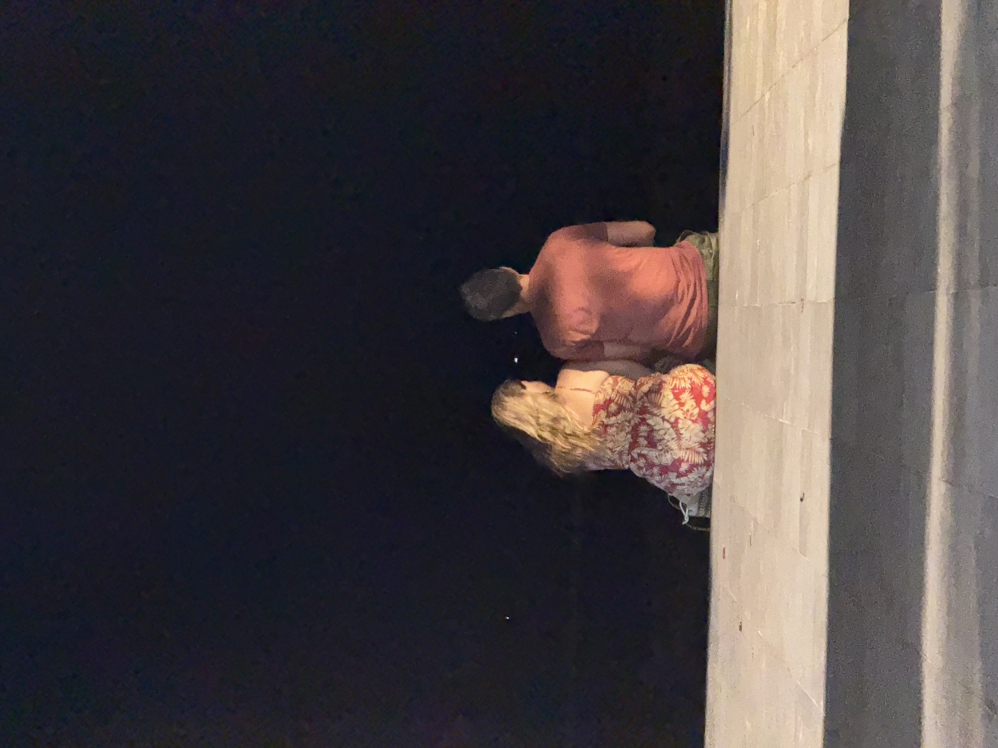 The image shows a couple sitting on a paved walkway at night, facing away from the camera and looking out at a dark expanse, possibly the ocean or a body of water. The woman is wearing a red and white floral dress, and the man is wearing a red t-shirt and camouflage shorts. The walkway appears to be made of large, light-colored rectangular tiles. The background is completely dark, with a few faint lights visible in the distance. The lighting suggests it is nighttime. This scene could be located along the coast in Denpasar, Indonesia, where such promenades are common.