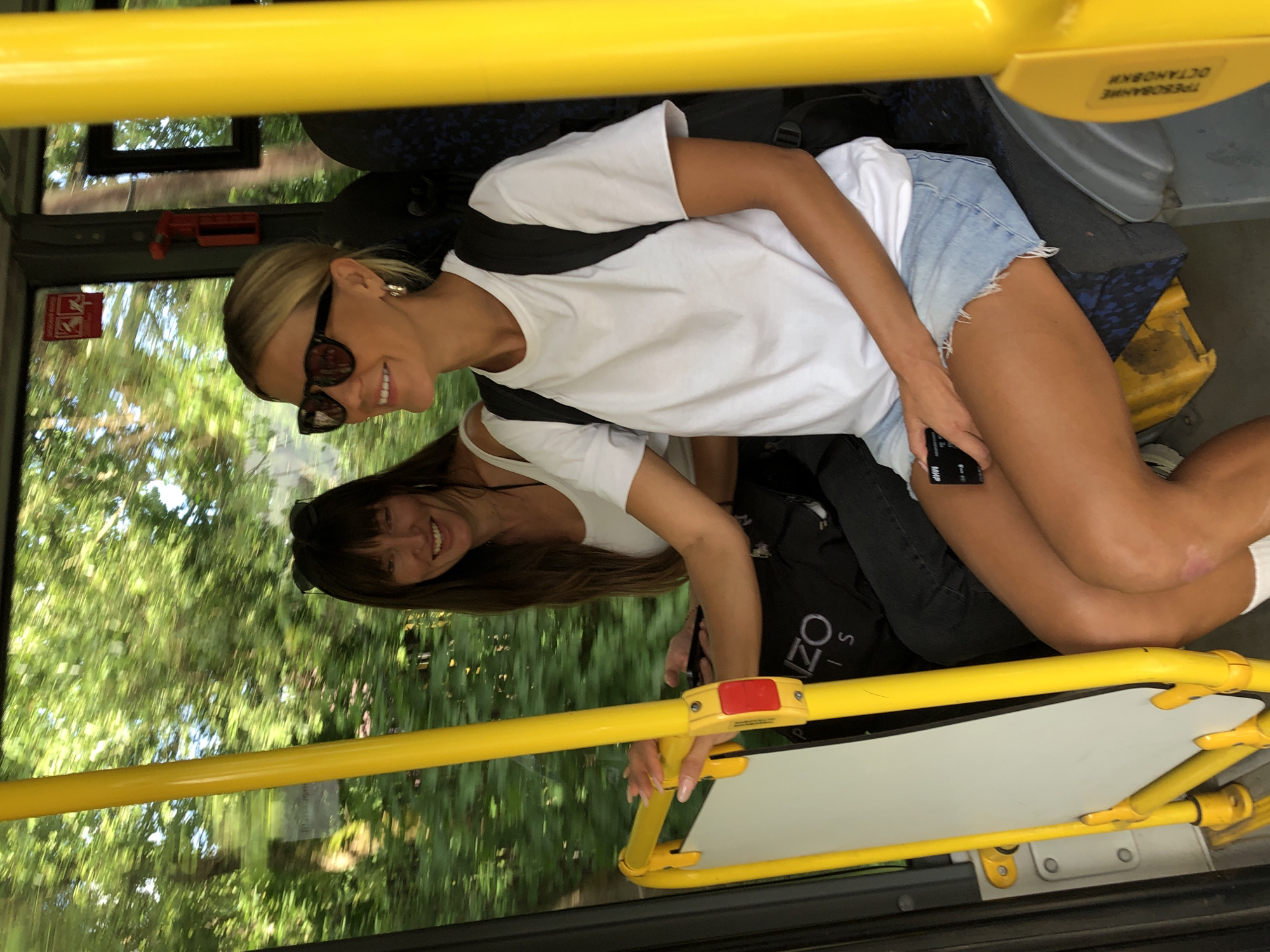 Two women are sitting on a bus, looking forward. The woman in the foreground is wearing sunglasses, a white t-shirt, and denim shorts. She is holding a phone in her right hand and has a backpack on her shoulders. The woman behind her is wearing a white top and has sunglasses on her head. The bus appears to be in motion, with trees visible through the windows. A yellow pole is to the right of the foreground woman. The bus seats are dark blue with a patterned design. A sign with Russian text is visible on the yellow pole on the right.
