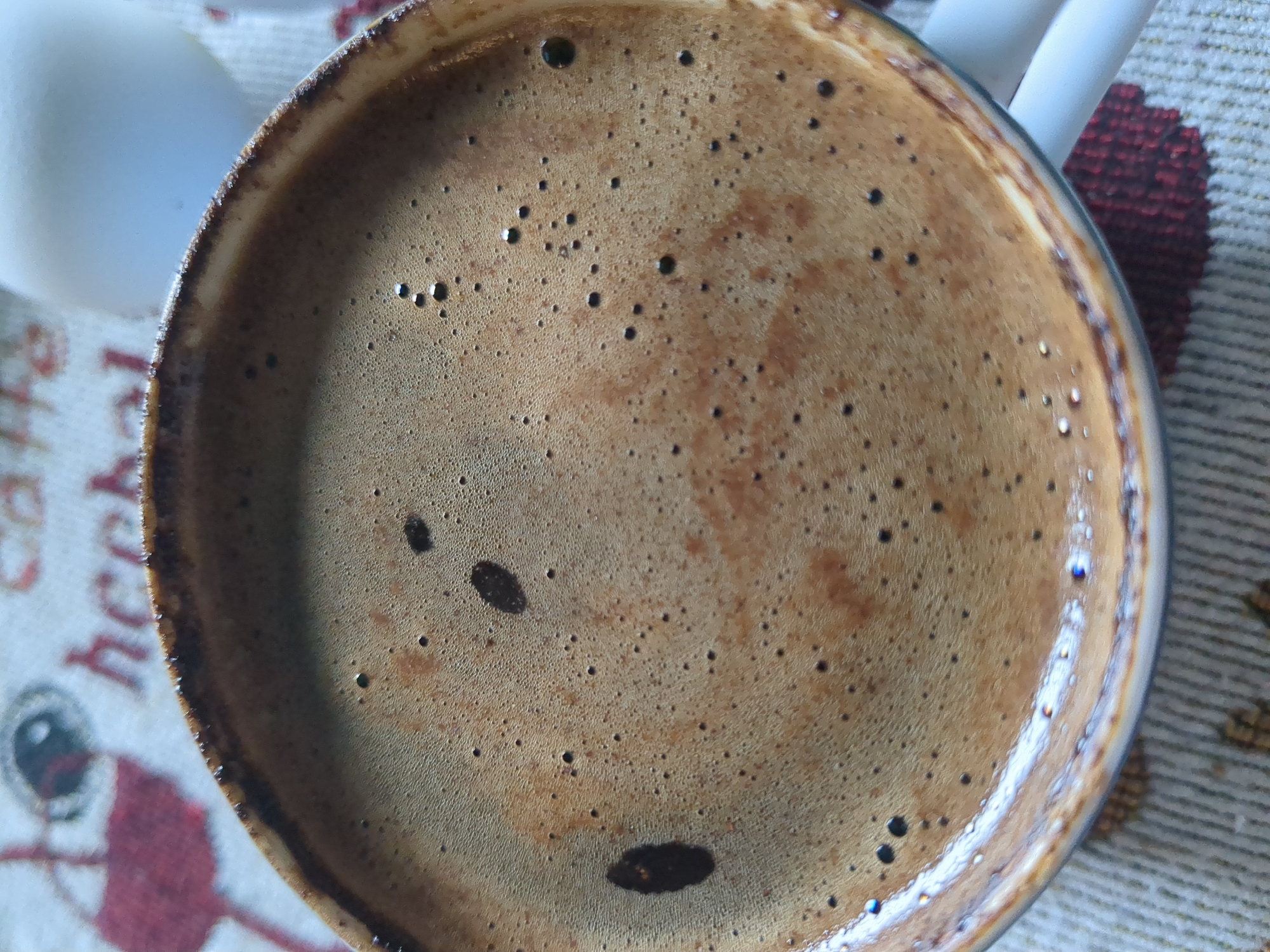 The image is a close-up, overhead shot of a cup filled with frothy coffee. The coffee has a rich brown color with many small bubbles forming the foam. There are a few larger dark spots within the foam, possibly from the brewing process or sugar. The rim of the cup is visible, appearing to be dark ceramic with a slightly rough texture. The cup is resting on a patterned tablecloth with visible red and white elements. In the background, out of focus, a white object, likely another cup or container, is partially visible. The lighting suggests an indoor setting. There is no indication of the city of Nordhausen, Germany in the image itself.
