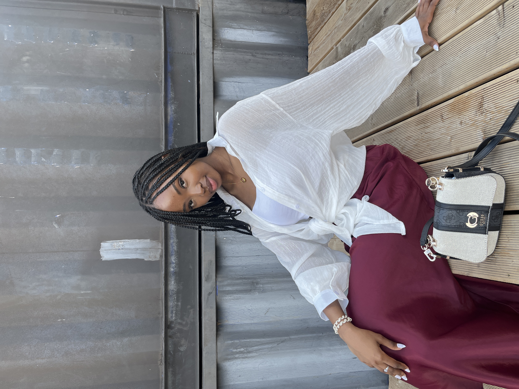 A young Black woman with braided hair is sitting on a wooden deck in front of a large, weathered metal structure. She is wearing a white, open, long-sleeved blouse tied at the waist, revealing a white crop top underneath. Her lower half is clad in a flowing, deep red skirt. She is accessorized with a white beaded bracelet on her left wrist, a ring on her left index finger, and a delicate gold necklace. A small, two-toned handbag with a prominent "GUESS" logo sits on the deck beside her. The woman is looking towards the camera with a slight smile. The background appears to be an urban industrial setting. There is no identifiable information to suggest the location is the city of Kazan, Russia.