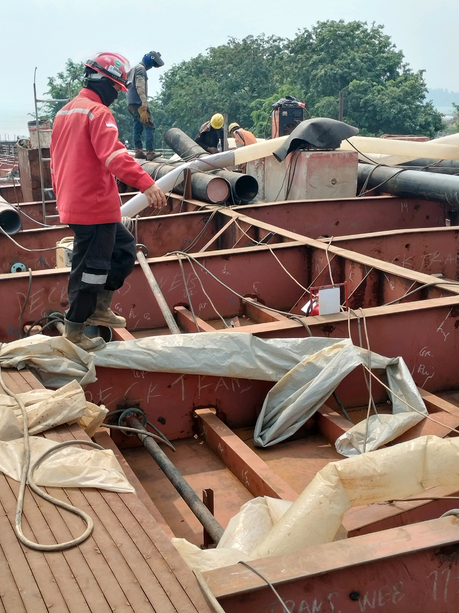 In Batam, Indonesia, a construction site features workers on a large-scale industrial project, likely shipbuilding or repair, evidenced by the substantial metal structures and pipes. In the foreground, a worker in a red jacket and hard hat stands on a wooden deck, holding a white pipe. Another worker, wearing a welding mask, is visible in the background, along with several other individuals in work attire scattered among the equipment. The environment is characterized by exposed metal beams, large pipes, welding equipment, and construction materials, suggesting an active and ongoing industrial operation. The weather appears overcast.