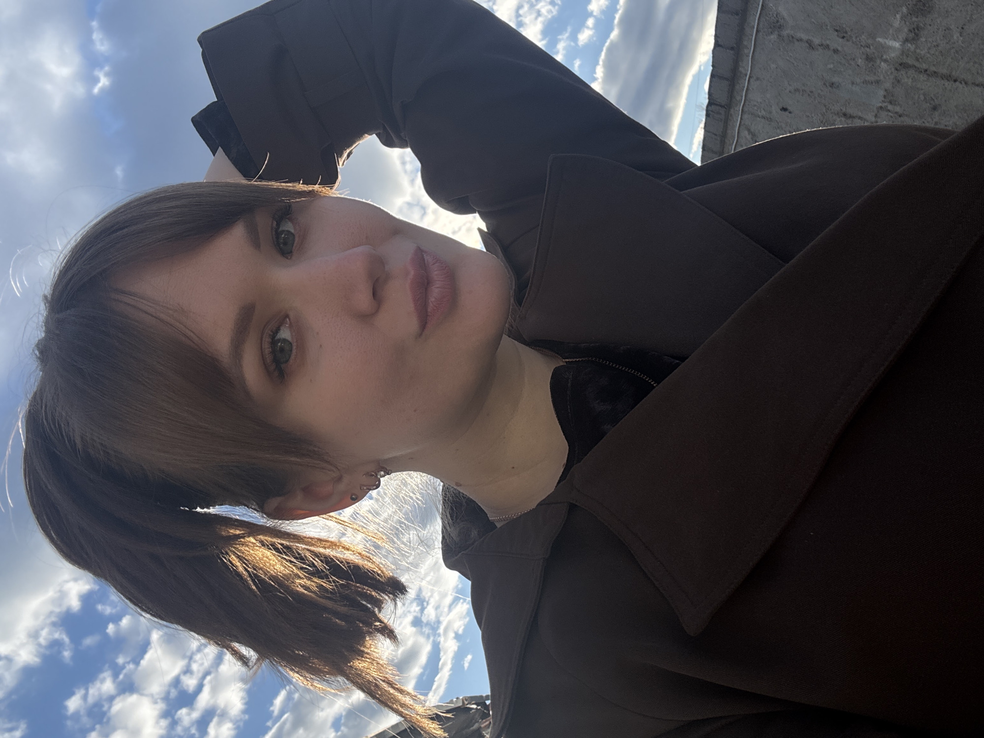 A young woman with fair skin and brown hair pulled back into a low ponytail stands outdoors, likely on a rooftop in Chaikovsky, Russia. Her brown bangs are visible, framing her face, and she has multiple piercings in her left ear. She is wearing a dark brown coat or jacket. The background features a bright, cloudy sky and the top edge of a concrete structure, potentially part of a building's roof or parapet. The image is taken from a low angle, looking upwards towards the woman.