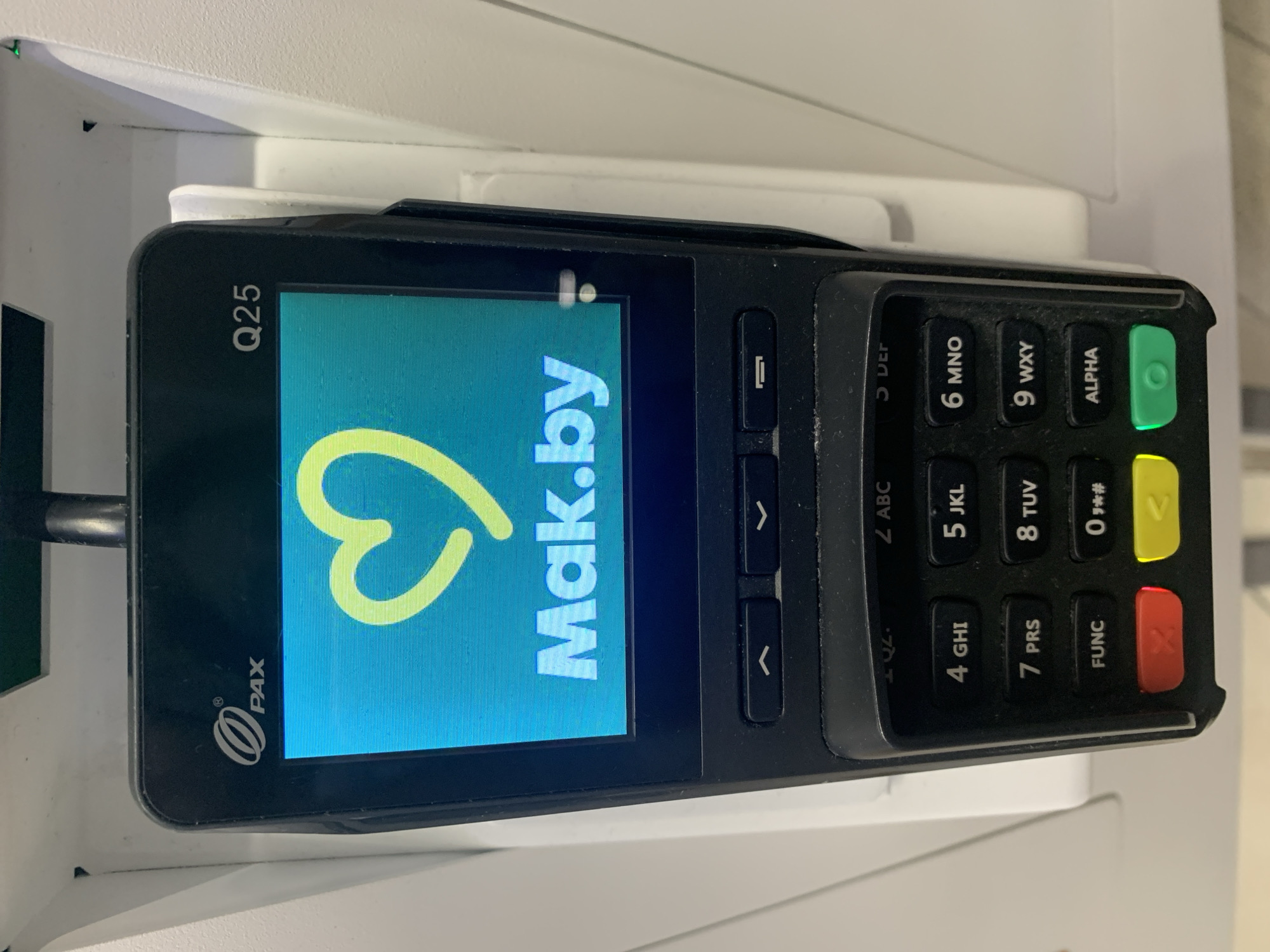 The image shows a close-up of a black and white PAX Q25 credit card terminal. The screen displays a light blue background with a large, yellow heart icon and the text "Mak.by" in white letters. Below the screen are several function buttons and a numeric keypad. The terminal is mounted on a white surface, likely a counter or stand, in what appears to be a public indoor space. There are no people visible in the image. The lighting is bright, suggesting an indoor environment with artificial or natural light. The perspective is from slightly above and to the side of the terminal. The terminal is clean but shows some dust accumulation on the keypad. The context of Hrodna, Belarus is not directly inferable from the image itself, but this type of terminal is commonly found in retail environments in that region.