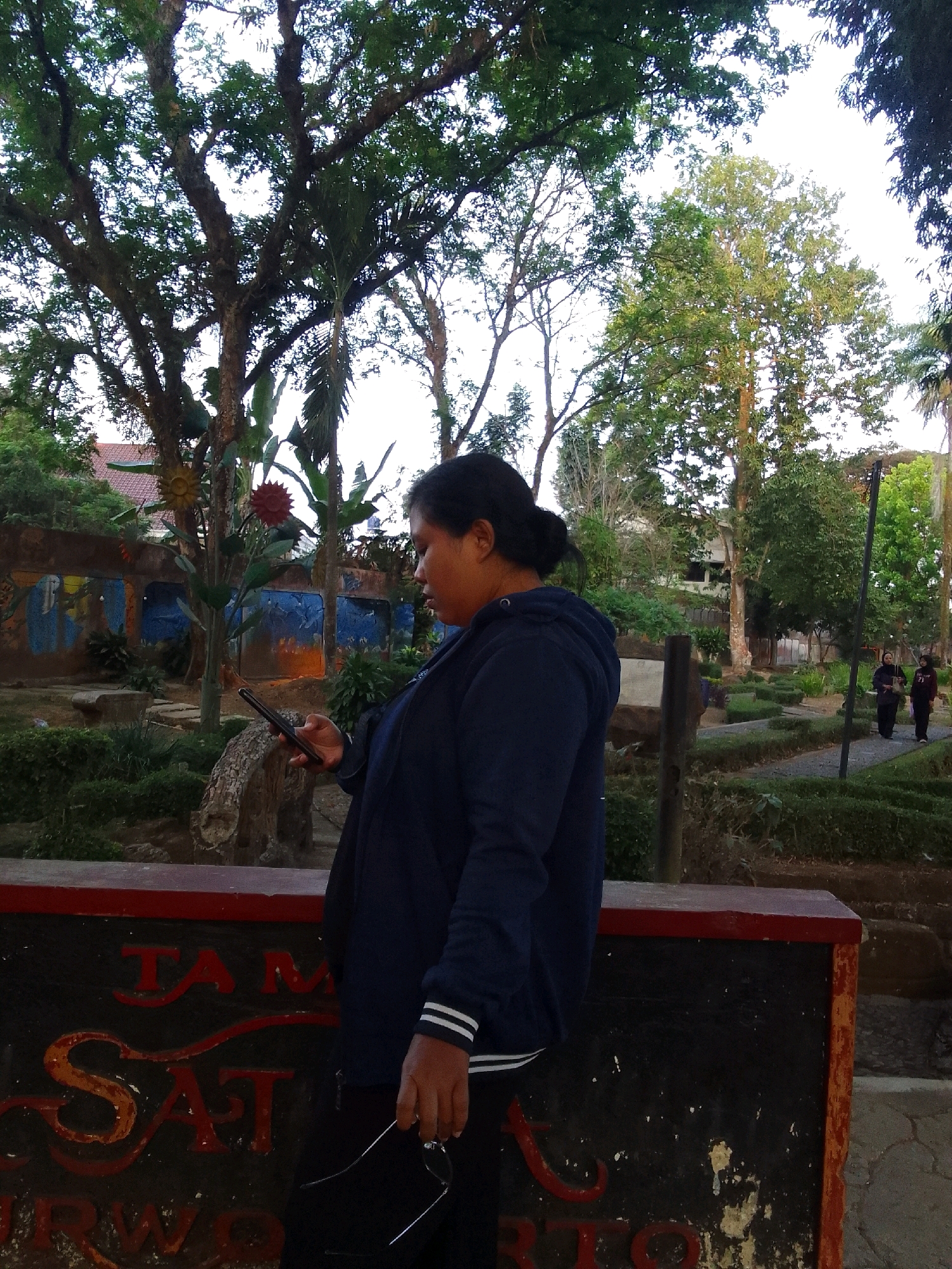 A young woman with dark hair tied back stands in a park or garden area, looking down at her black smartphone. She is wearing a dark blue hooded sweatshirt with white stripes on the cuffs and black pants. She holds a pair of glasses in her right hand. Behind her, a low wall with red and black paint is visible, featuring red lettering. Beyond the wall, there are trees, foliage, and a path with manicured bushes. In the background, two women in dark clothing are walking along the path. The scene appears to be in a public outdoor space, possibly in Desa Pamijen, Kecamatan Sokaraja, Indonesia, given the style of vegetation and architecture. The lighting suggests it is daytime, possibly late afternoon or early morning.