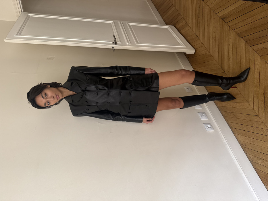 An individual stands in an interior room, characteristic of a Parisian apartment, evidenced by the herringbone parquet flooring and paneled white door. The subject is a young adult female with short, dark hair, positioned centrally, looking directly at the viewer with a neutral expression. She is wearing a black, double-breasted leather blazer-style mini-dress or jacket featuring a notched lapel, multiple buttons, and four pockets (two chest, two hip). Her lower legs are bare, ending in knee-high, black, pointed-toe stiletto boots; the right boot displays a distinct red sole. The background consists of a plain, light-colored wall with minor blemishes, a white baseboard, and several white electrical outlets. To the right, a tall, white, paneled interior door with a silver handle is partially ajar.