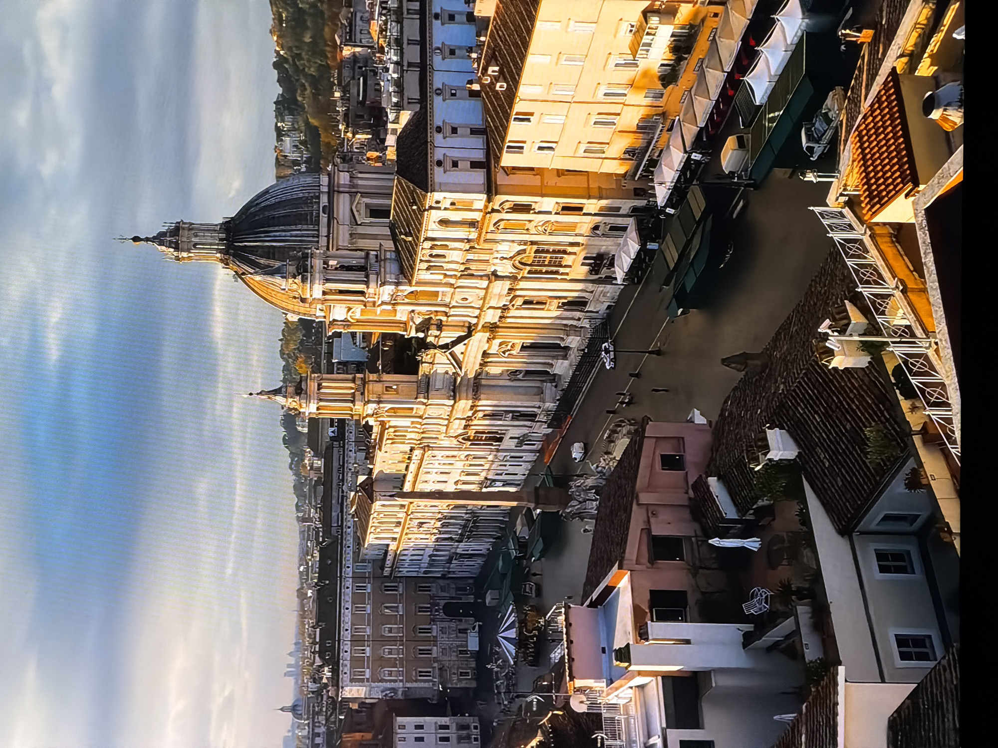 The image shows an aerial view of Piazza Navona in Rome, Italy, bathed in the warm light of sunrise or sunset. The prominent Church of Sant'Agnese in Agone, with its Baroque facade and dome, dominates the right side of the frame. In front of it, the Fountain of Four Rivers, sculpted by Gian Lorenzo Bernini, is visible as a collection of white marble figures. An Egyptian obelisk stands near the fountain.
The piazza itself appears to be set up as a market, with numerous stalls covered by green and white awnings lining the street. Several vehicles, including vans and what appears to be a truck, are parked or driving through the area. In the center of the piazza, a carousel with colorful decorations is set up, suggesting a festive atmosphere.
The surrounding buildings are a mix of architectural styles, with many featuring terracotta tiled roofs. The sky above is dramatic, with a mix of blue and grey clouds, some illuminated by the low-angled sun. The overall impression is one of a historic European city square coming to life with a morning market and recreational activities.