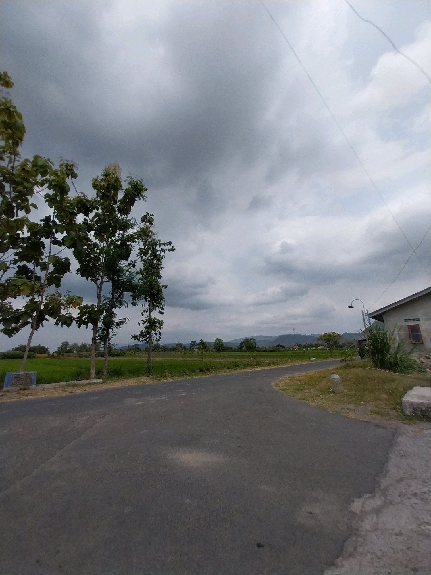 The image depicts a rural scene in Cucukan, Indonesia, under a cloudy sky. A paved road curves from the foreground into the middle ground, bordered on the left by lush green rice fields and several trees. On the right side of the road, a simple house with a reddish-brown window and a light fixture extends into the frame. The environment appears calm and natural, with a few scattered objects like a plastic bottle and a concrete block near the road.