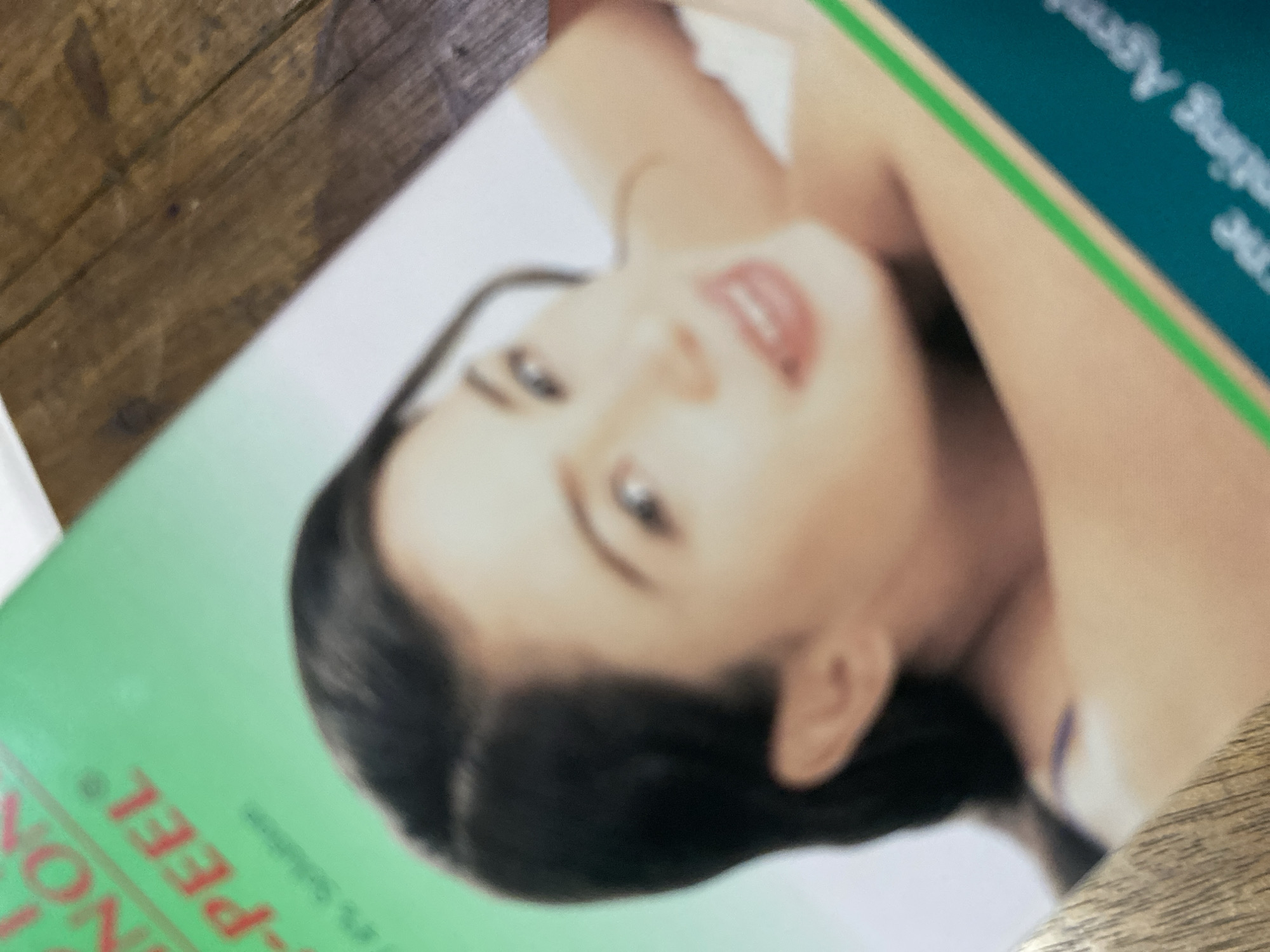 The image shows a close-up of a magazine or brochure featuring a woman's face. The woman has dark hair, fair skin, and is looking slightly upwards with a gentle smile. There is text on the left side of the image, partially obscured, with the words "NON-PEEL" visible in red. On the bottom right, there is text with a green stripe, and the words "nting Agent" are partially visible. The background appears to be a wooden surface. The context of Ta', Indonesia is not present in the image.