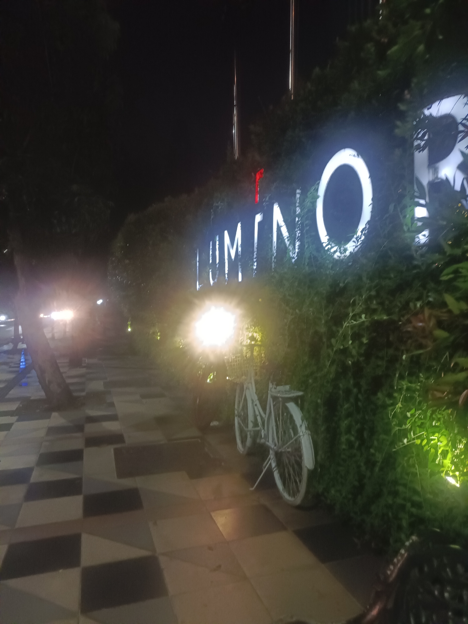 At night in Unknown, Unknown, a photograph shows a tiled walkway with an illuminated sign reading "LUMINO" surrounded by dense greenery. A white bicycle leans against the greenery near the sign. The walkway leads into the darkness, with faint lights visible in the distance. A tree stands on the left side of the walkway. The scene appears to be a public area, possibly a park or outdoor dining area.