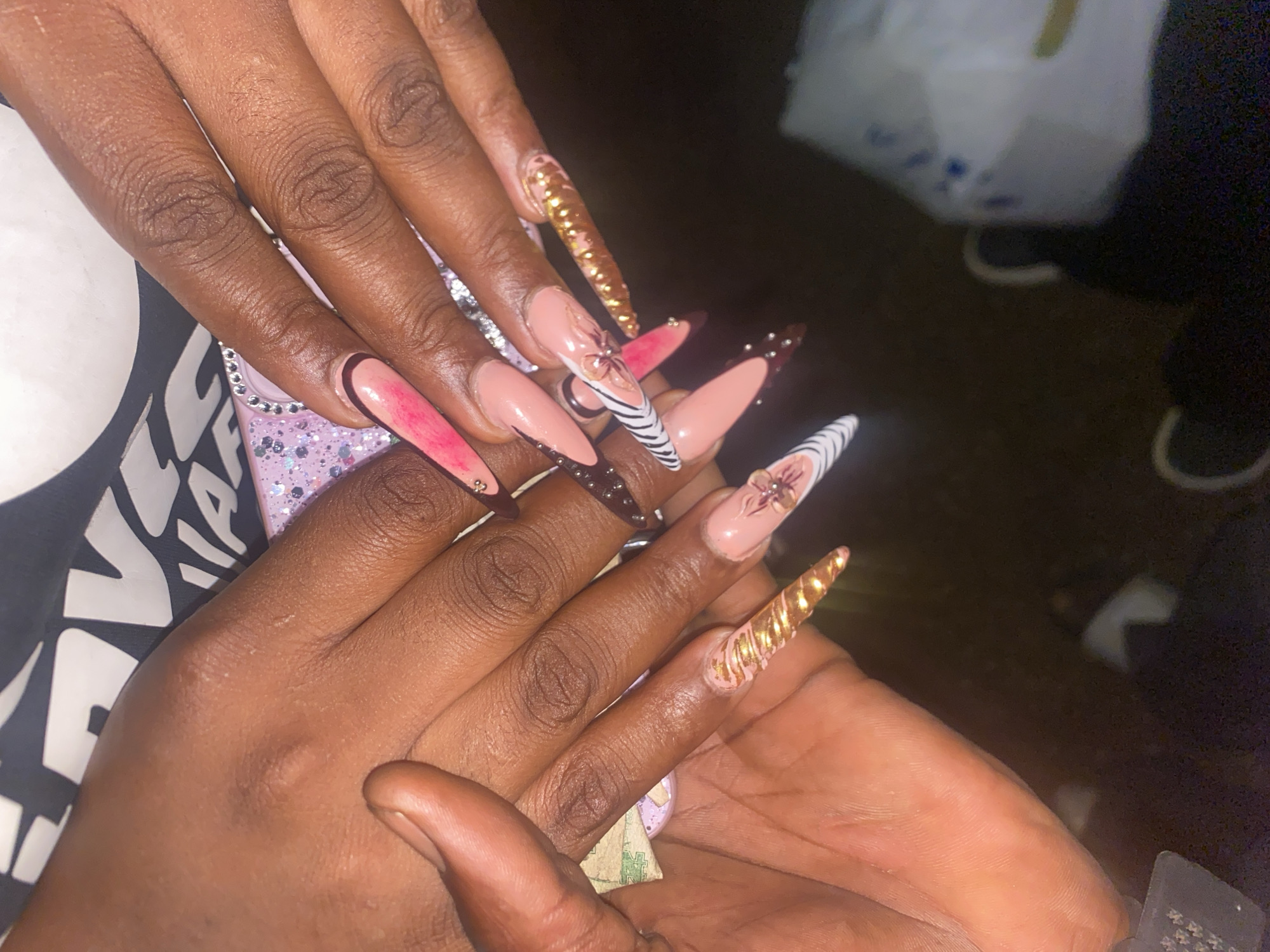 A pair of dark-skinned hands is displayed, showcasing several long, pointed artificial nails. The nails are adorned with various designs: one nail features a pink and black outline, another has a zebra-striped pattern with a flower, one is gold and spiraled, another is embellished with gold glitter, and yet another is colored in brown with dark brown dots. One hand holds a purple phone case, while the other appears to hold a folded US dollar bill. The setting appears to be indoors, with a dark background and a partially visible white bag in the upper background. There is no specific evidence in the image to confirm that the image was taken in Bukuru, Nigeria.