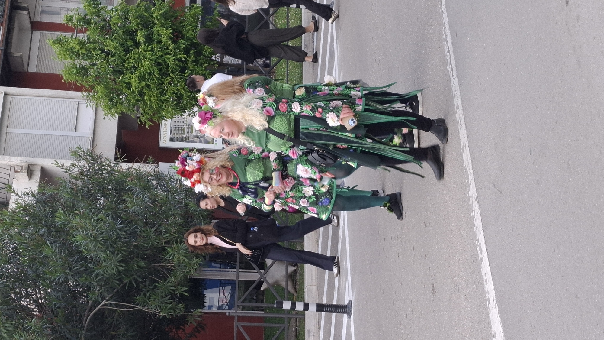 Two women dressed in floral costumes with flower crowns stand on a street near a building in Budva, Montenegro. They are surrounded by other individuals, some in casual attire. A row of trees lines the edge of the street beside the building. A black-and-white striped bollard is near the curb. The setting appears to be an outdoor urban environment during daylight.