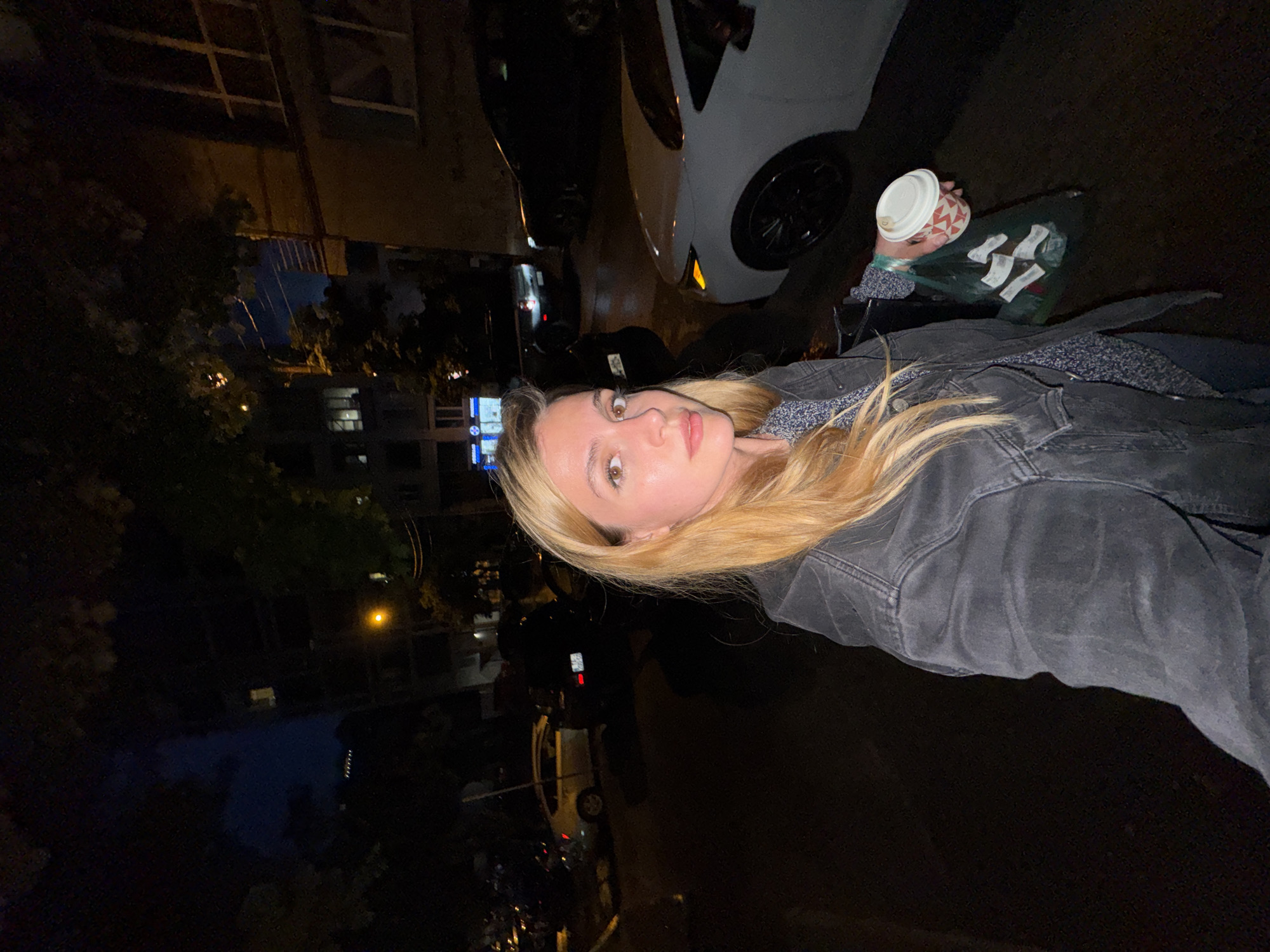 The image, rotated 90 degrees counter-clockwise, captures a young woman posing for a photo at night in an urban setting, likely in Tbilisi, Georgia.
The central subject is a woman with long, straight blonde hair and light brown eyes, looking directly at the camera with a neutral expression. She wears a dark, possibly charcoal grey, denim jacket over a patterned dark top. In her right hand, she holds a disposable coffee cup with a white lid and a red and white geometric design. Below the cup, she carries a dark green reusable bag, containing several individually wrapped items in light-colored packaging, though any text on them is illegible.
The background is dimly lit, indicating nighttime. A white car with a black wheel is parked behind her. Further back, dark vehicles and the silhouettes of buildings are visible, some with illuminated windows. Dark foliage from trees frames parts of the scene. A solitary yellow streetlight casts a warm glow in the middle-right background, contributing to the evening ambiance of the city street or alleyway. The overall scene suggests a casual moment during an evening outing in an urban environment.