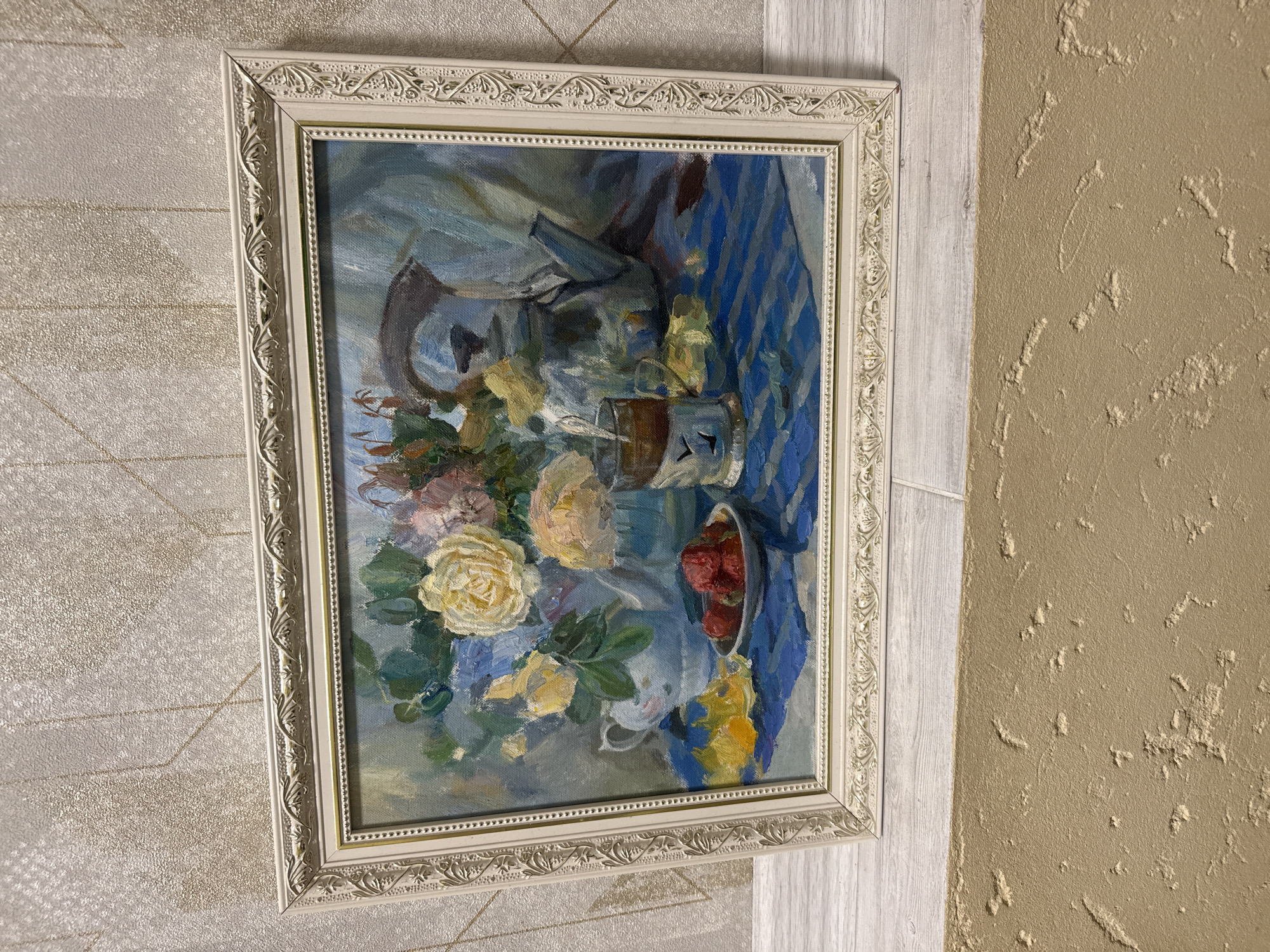 A framed painting hangs on a wall. The painting is a still life depicting flowers, fruit, and glassware on a blue and white striped cloth. The frame is ornate and white. The wall has two distinct textures, one a textured beige and the other a textured off-white. The location is unknown.