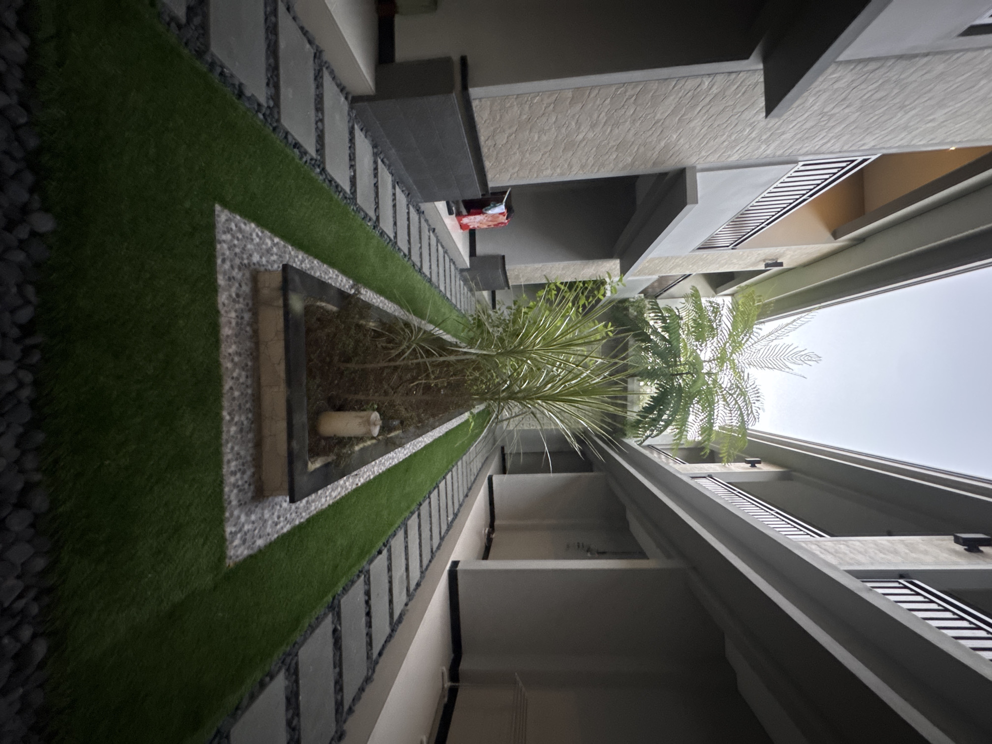 The image shows a narrow outdoor walkway between two modern buildings. The walkway is paved with large, dark square stones interspersed with dark pebbles, and a strip of artificial grass runs alongside it. In the center of the walkway, a raised planter box contains several plants, including a fern and a tall, spiky plant. The buildings have balconies with dark railings and the exterior walls have a textured, light-colored finish. Clothes are hanging on a rack on the left side of the walkway, near an open doorway. The scene appears to be in a contemporary residential or hotel complex in Kuta, Indonesia.