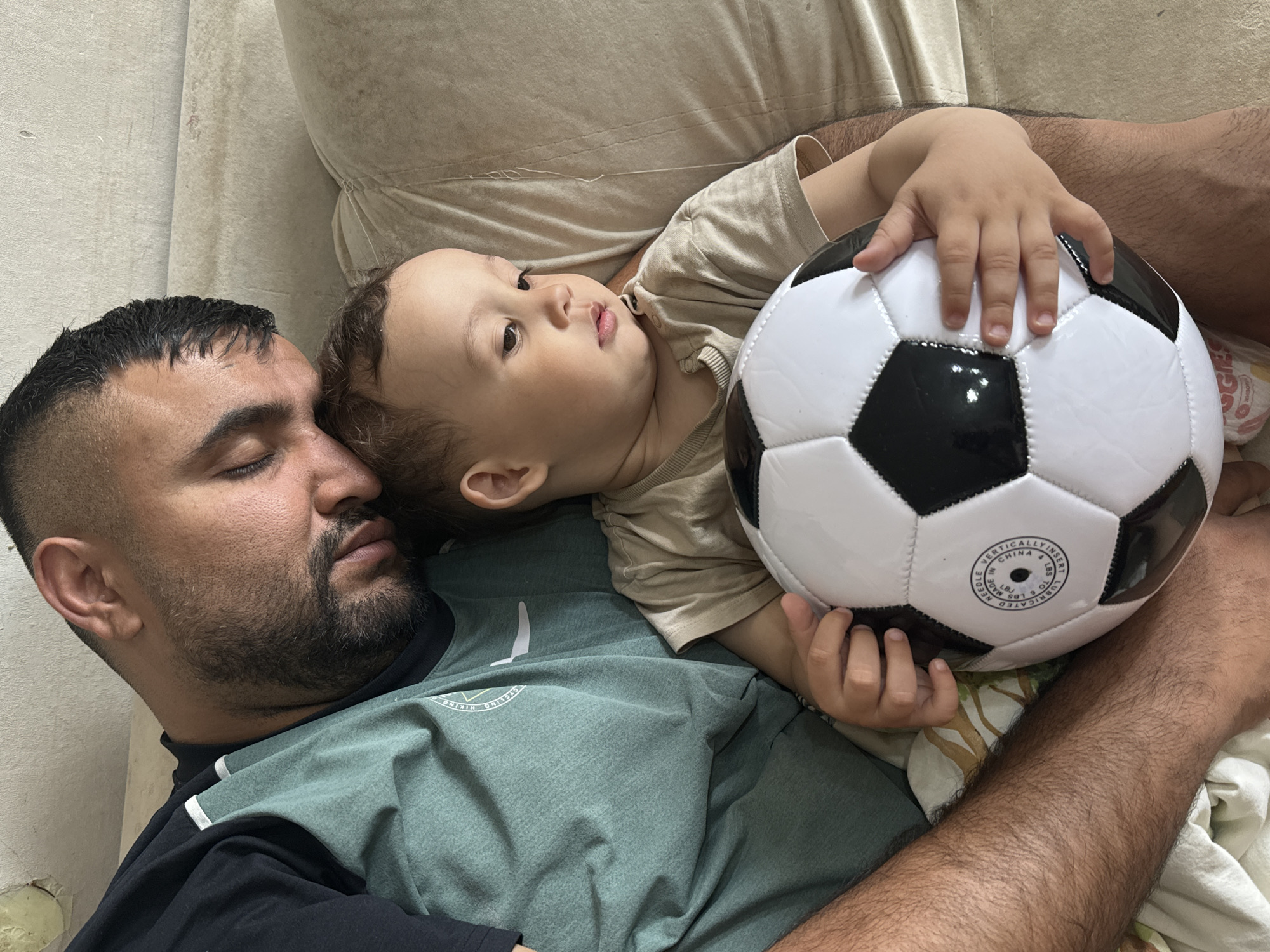 The image depicts a man and a young child lying down together. The man has short dark hair, a beard, and is wearing a two-tone green and black shirt. He appears to be sleeping. The child, lying on the man's chest, is wearing a light brown shirt and holding a black and white soccer ball. The background suggests an indoor setting, possibly a living room or bedroom. The location is not specified in the image, and therefore the city of Chaikovsky, Russia cannot be confirmed.