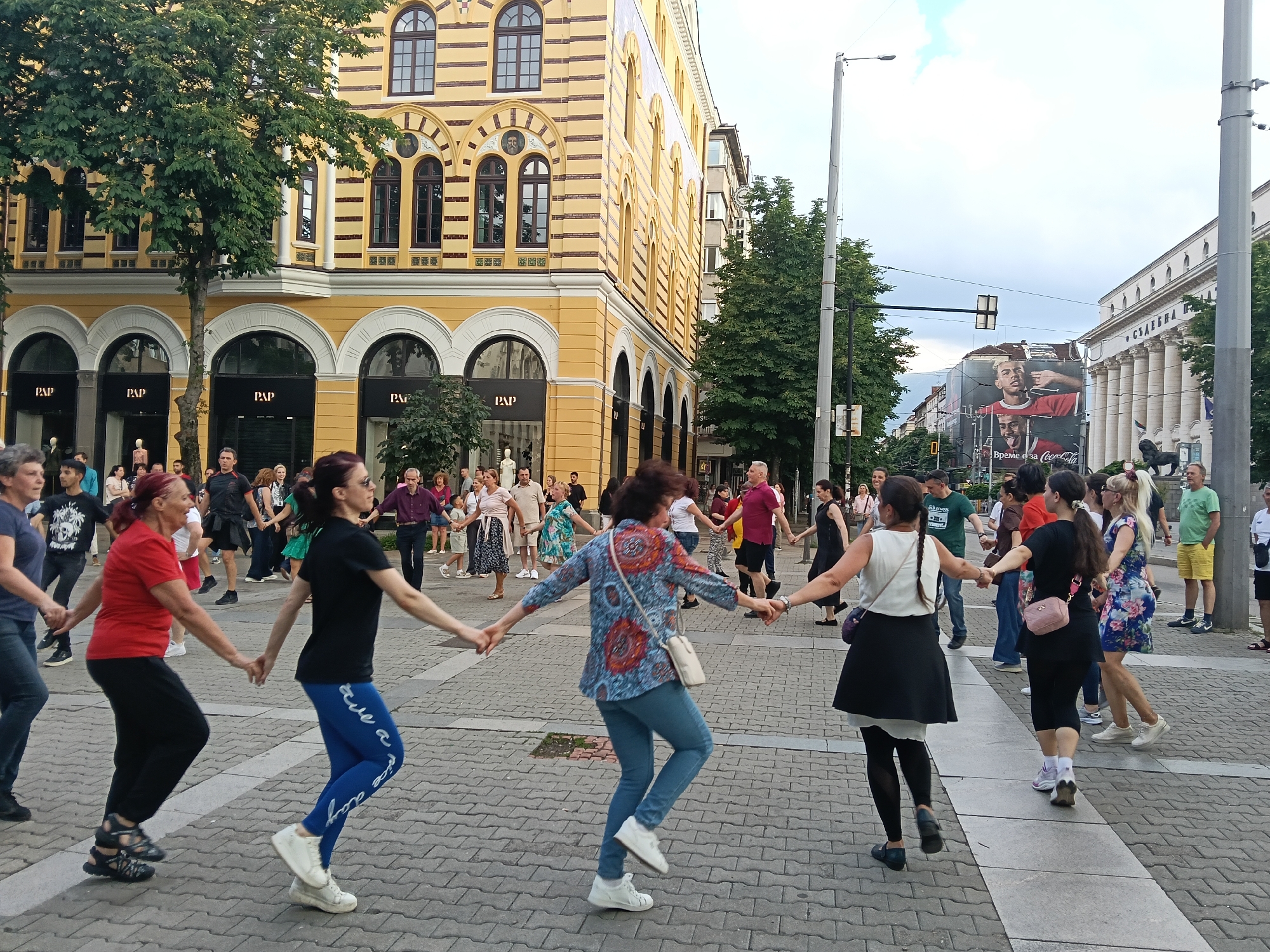 The image depicts a large group of people engaged in a traditional folk dance (Horo) in a central pedestrian square. The location is Knyaz Alexander I Square (also known as Battenberg Square) in Sofia, Bulgaria.
Approximately 30-40 individuals, ranging in age from young adults to seniors, are holding hands in a large, undulating line or semi-circle, moving rhythmically across the paved square. They are dressed in diverse casual modern attire, with various colors and styles visible. Their body language and foot positions indicate coordinated dance steps.
In the background to the right, the prominent neoclassical facade of the National Assembly of Bulgaria (Parliament) building is visible. It features tall columns, a pediment, and a large flight of steps. A dark-colored lion statue is discernible at the base of the building's steps. Further right and behind the Parliament, a large billboard displays a Coca-Cola advertisement featuring two men, with Cyrillic text "ВРЕМЕ Е ЗА Coca-Cola" (Time for Coca-Cola).
To the left and center of the frame, a multi-story building with a distinctive yellow and brown striped facade stands. This building exhibits numerous arched windows on its upper floors and several arched storefronts at ground level, some displaying "PAP" signage. A mature tree with green foliage stands in front of this building.
The ground is paved with interlocking pavers in various shades of grey and reddish-brown, arranged in geometric patterns. Streetlights and other smaller trees are visible in the mid-ground. The sky is overcast, indicating either late afternoon or early evening light conditions.