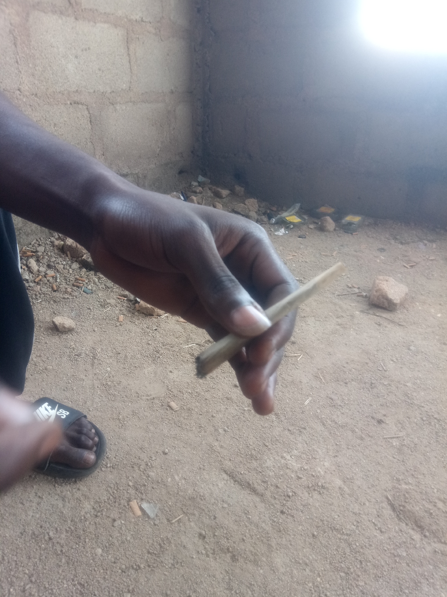 An adult's hand, with dark skin, holds a small, light-colored cigarette or rolled item, possibly a joint. The hand is positioned in the lower right quadrant of the image, with the item held between the thumb and forefinger. The person is wearing a black slide sandal with a white logo on the left foot, visible in the lower left corner. The background consists of a dirt ground with scattered debris, including cigarette butts, small rocks, and some yellow-wrapped items. A rough, concrete brick wall is visible in the upper part of the image. The lighting suggests an outdoor or semi-enclosed environment, possibly during the day due to the bright glare in the upper right corner.
