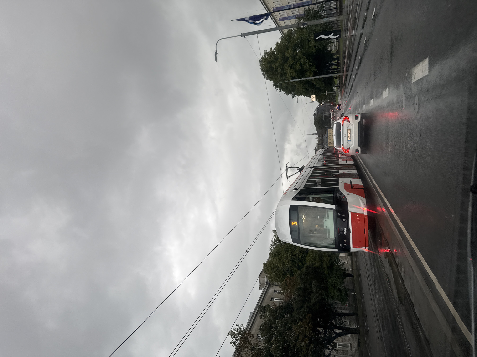 A modern white and red tram numbered '3' is the primary subject in this slightly overcast, rainy scene in Tallinn, Estonia. The tram is on a wet road, with visible reflections on the asphalt. A white car, a Honda Civic with license plate '481 MSB', is driving behind the tram. In the background, there are trees, buildings with a classical architectural style, and streetlights. The sky is filled with gray, cloudy formations, typical of a rainy day. Electrical wires for the tram are visible overhead. The overall impression is a wet, urban environment in a European city.