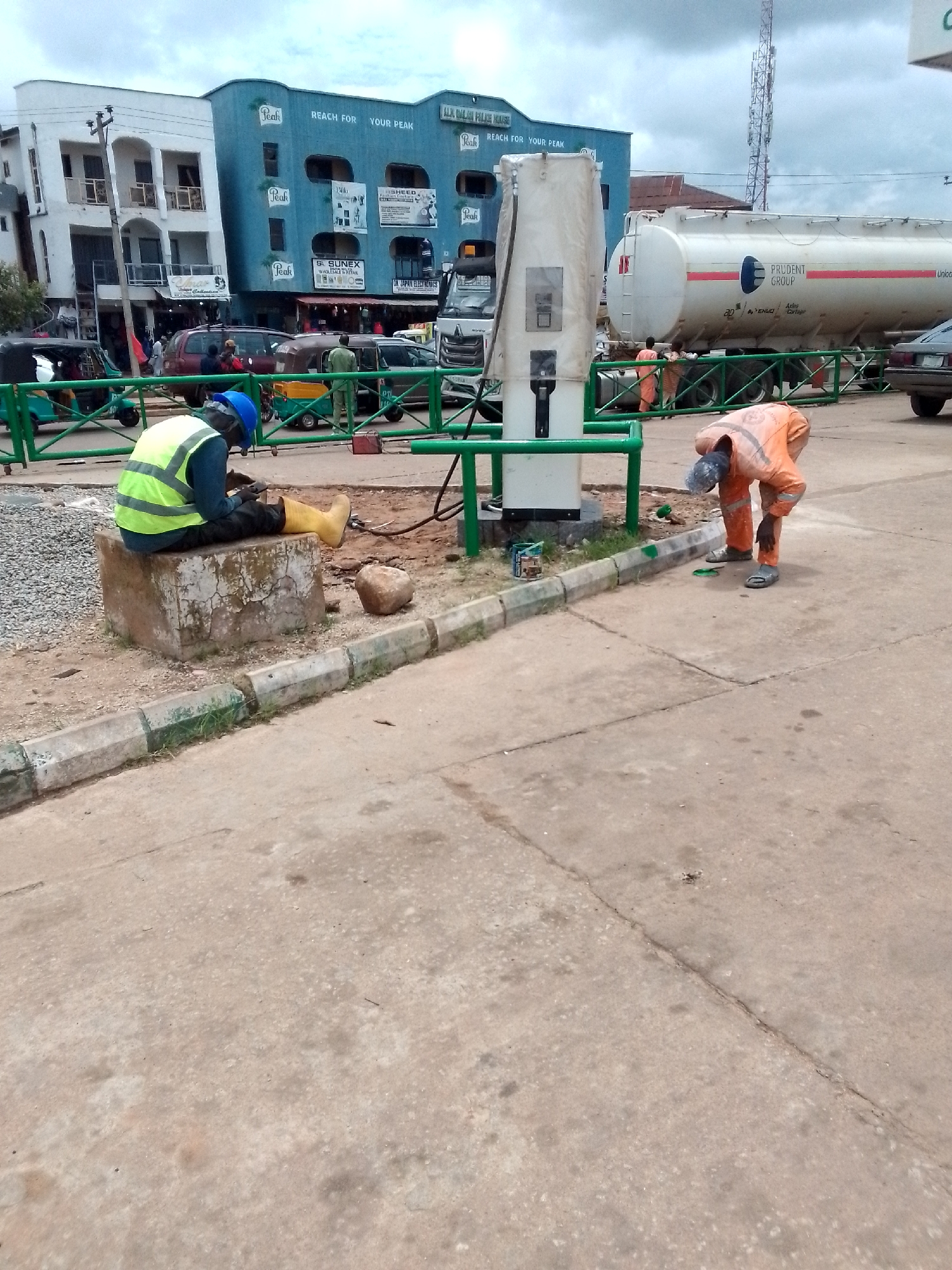 In Jos, Nigeria, two construction workers, one in a bright yellow vest and blue hard hat, and another in an orange jumpsuit, are seen working near a large white fuel tanker truck and a gas pump. The worker in the yellow vest is seated on a concrete block, appearing to be taking a break or working on something in his lap. The worker in the orange jumpsuit is bent over, possibly cleaning or performing maintenance on the ground near the gas pump. In the background, a multi-story building with "Peak" branding is visible, along with other vehicles and a cellular tower. The scene suggests an active construction or maintenance site in an urban environment.