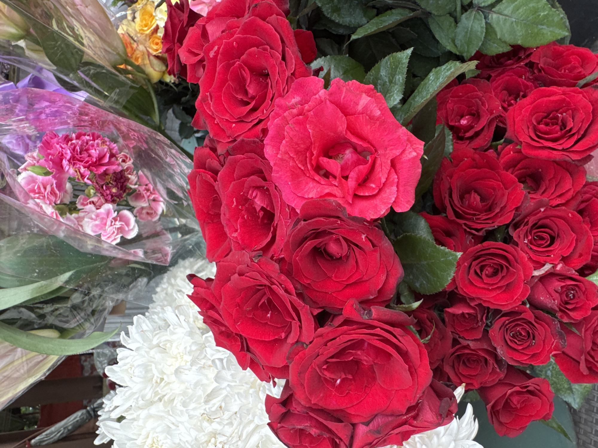 The image shows a close-up view of a bouquet of flowers, predominantly red roses. There are also white chrysanthemums and pink carnations visible in the background. The flowers appear to be fresh and are arranged in a bouquet. The lighting suggests an outdoor setting, possibly a flower shop or market. The scene is static, with no people or specific actions depicted. The environment appears to be a retail display for flowers. The provided crops focus on different sections of the bouquet, highlighting the textures and colors of the various flowers.