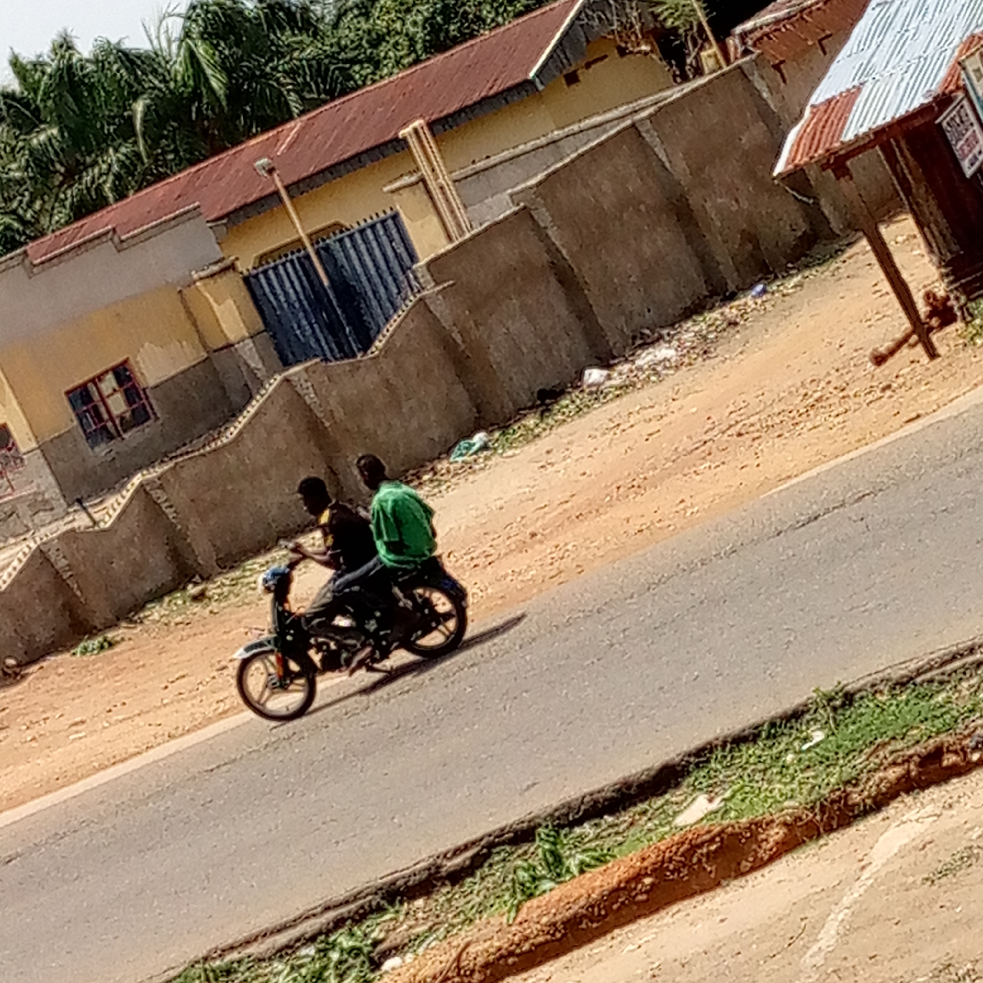 A medium shot depicts two individuals riding a motorcycle on a paved road in an urban environment. The foreground features a section of the road with grass and soil bordering it. To the left, there's a building with a tan exterior, a red-tiled roof, and a blue gate. A concrete retaining wall extends from the building, and the background reveals additional buildings and lush green trees. Debris is scattered on the ground near the wall.