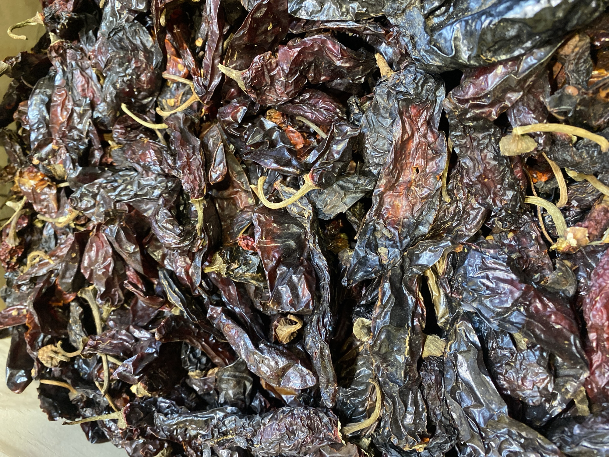 The image shows a close-up of a pile of dried chili peppers. The peppers are dark red and brown in color, and their skins are wrinkled and shiny. Some of the peppers have stems still attached. The photo appears to have been taken indoors. The location of the photo is San Bernardino Tlaxcalancingo, Mexico.