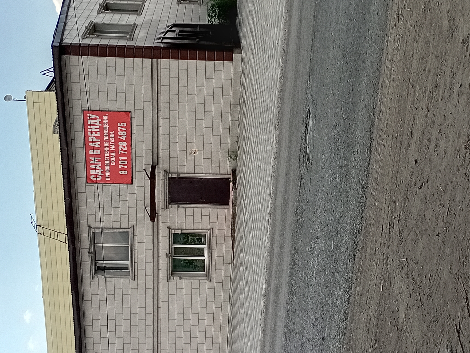 The image shows a two-story building with a light-colored brick facade. A large red sign with white lettering is prominently displayed on the front of the building. The sign reads "СДАМ В АРЕНДУ" at the top, followed by "ПРОИЗВОДСТВЕННОЕ ПОМЕЩЕНИЕ, СКЛАД, МАГАЗИН" in smaller text, and finally a phone number "8 701 728 4875".
The building has several windows, some with white frames and others with dark frames. There is a dark brown door on the first floor. A black metal gate is visible on the right side of the building. The roof is light-colored and appears to be made of metal sheeting, with a ladder attached to it. The building is situated on a street with a paved surface in the foreground and a dirt shoulder. There is a tiled walkway in front of the building. The sky above is blue with some clouds. The location is identified as Semey, Kazakhstan.