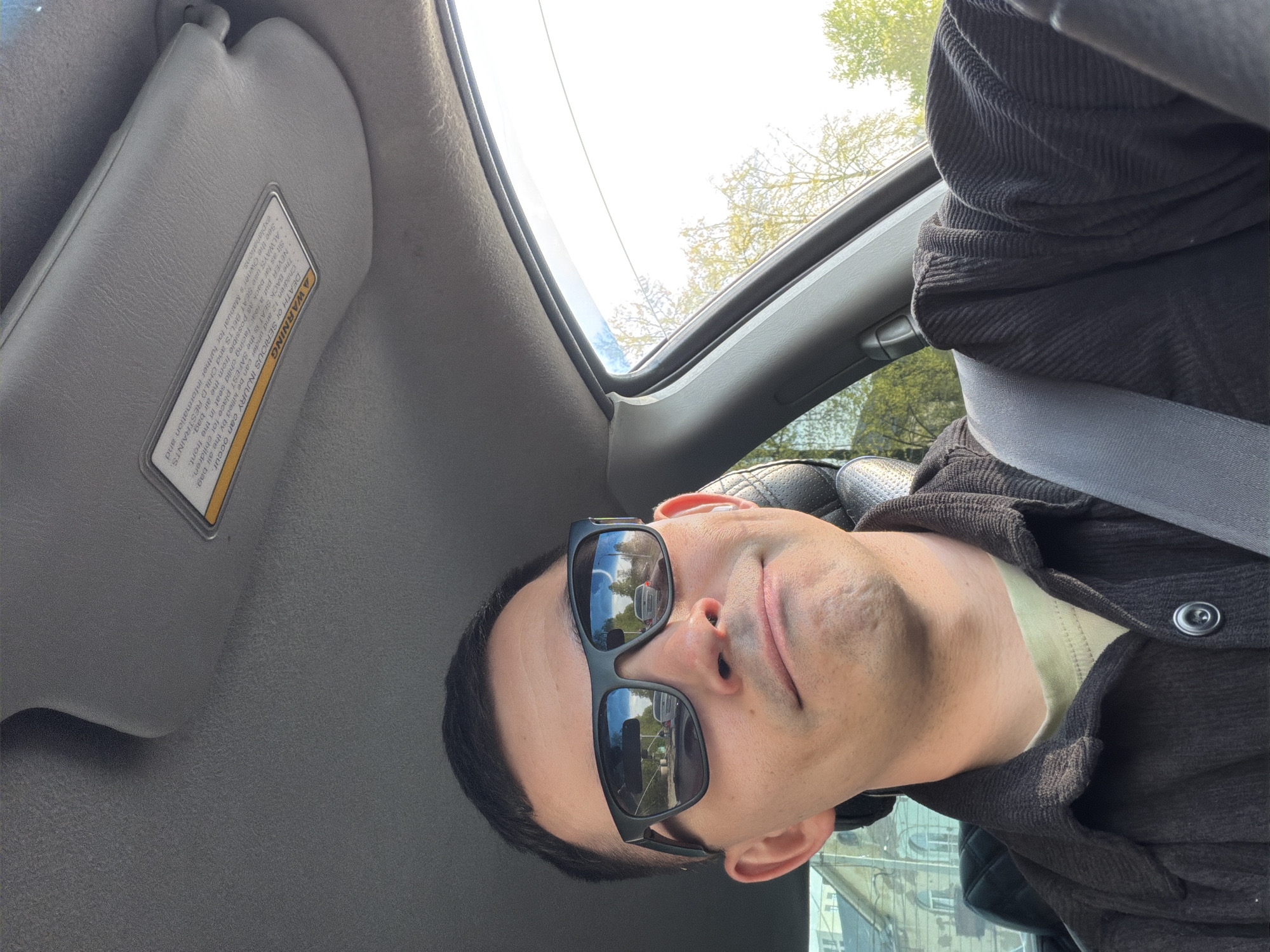 The image depicts a man wearing sunglasses and a dark button-down shirt in a car. He is wearing a seatbelt. The car has a sunroof and a sun visor. The reflections in his sunglasses suggest a city environment. Based on the location that you provided, the city environment in the sunglasses is Izhevsk, Russia.