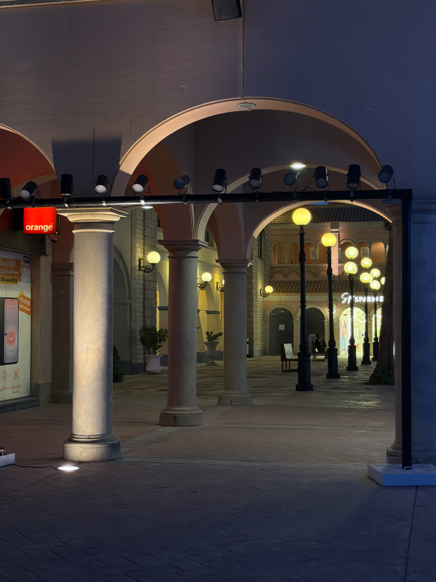 This is a nighttime photograph of an outdoor plaza in Sharm El Sheikh, Egypt, likely within a commercial or entertainment district. The scene is characterized by its architectural arches, stone columns, and multiple light sources. Prominently featured is an "orange" logo sign mounted on a red square, attached to the left of a large stone column. To the left of this column, a large advertisement for "Galaxy A37" is displayed on a screen or poster, showing a smartphone.
The plaza is paved with textured stone tiles. Several tall, black lampposts with round, illuminated globes line the walkway in the background, casting a warm glow. More lighting is present under an archway in the mid-ground, with spherical lamps attached to wall sconces. A structure with multiple spotlights directed downwards is visible spanning across the top of the frame, above the arches and columns, illuminating the walkway. In the distance, through an archway, a store named "SPARKLE" is visible, with illuminated signage. A single person is seated on a bench in the distance, observing the scene. The overall ambiance is one of evening illumination in a developed urban environment.