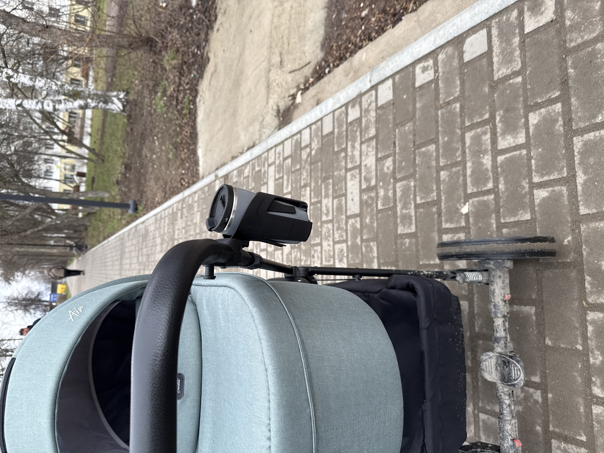 The image shows a first-person perspective from a stroller moving on a brick walkway. A camera is mounted on the stroller's handlebar. The stroller is positioned on a sidewalk, with a landscaped area featuring grass, trees, and a concrete path visible on the left side. A row of buildings lines the opposite side of the street. The stroller's frame, wheels, and the interior of the stroller seat are visible, suggesting a walk through an urban area.