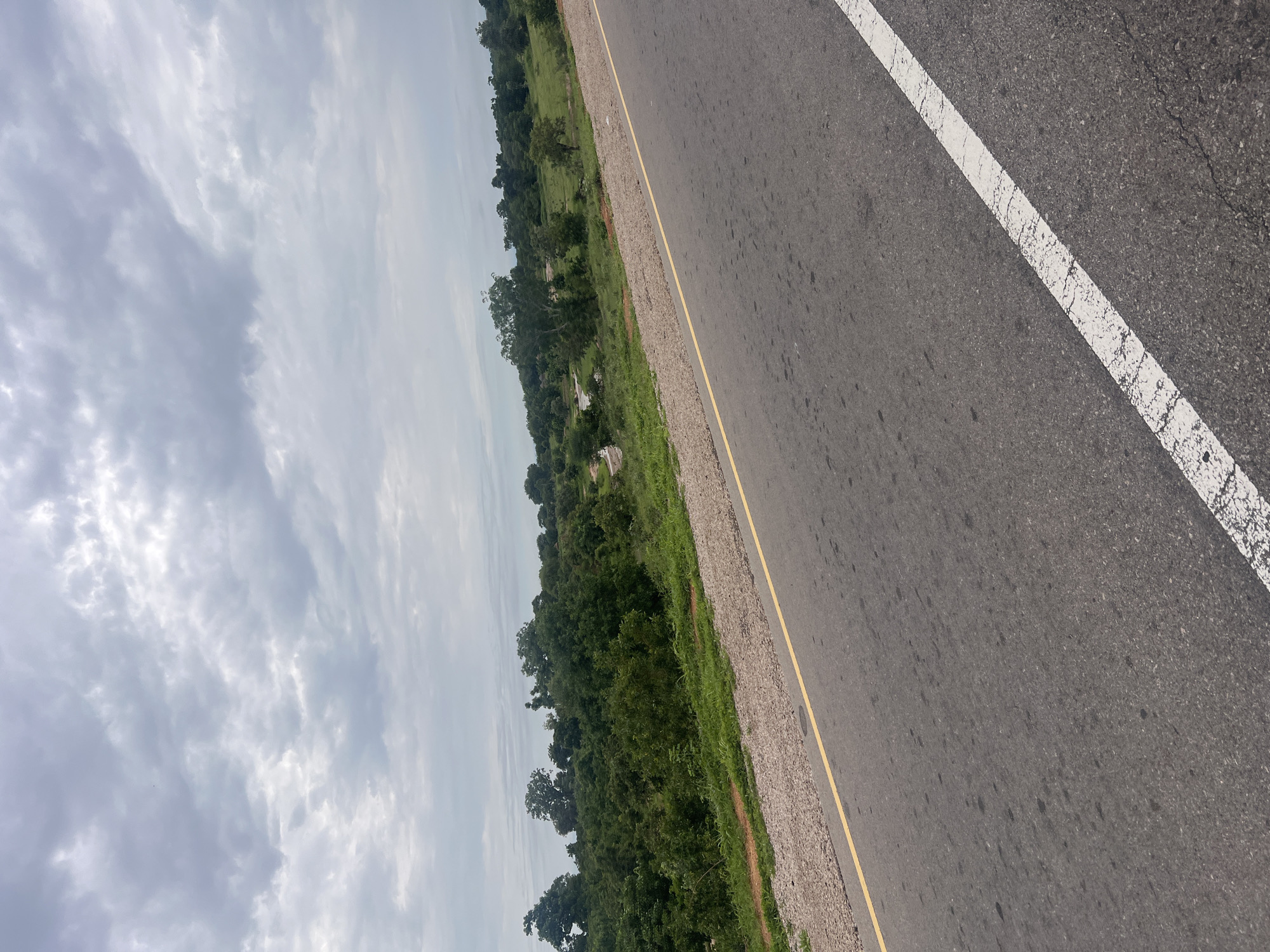 The image depicts a roadside scene in Minda, Nigeria. The perspective is from a moving vehicle, looking outwards. The foreground is dominated by an asphalt road with a white line marking its edge. A yellow line runs parallel to the white line, separating the road from a gravel shoulder. Beyond the shoulder, lush green vegetation and trees line the side of the road, suggesting a rural or semi-rural environment. The sky is overcast with dramatic, textured clouds, indicating a cloudy day. The lighting is diffused, and there are no clear shadows, further supporting the notion of overcast weather. The overall mood is neutral, with no discernible human activity or specific events captured. The image focuses on the landscape and the road, providing a sense of travel and the surrounding environment. No text is visible in the image.
