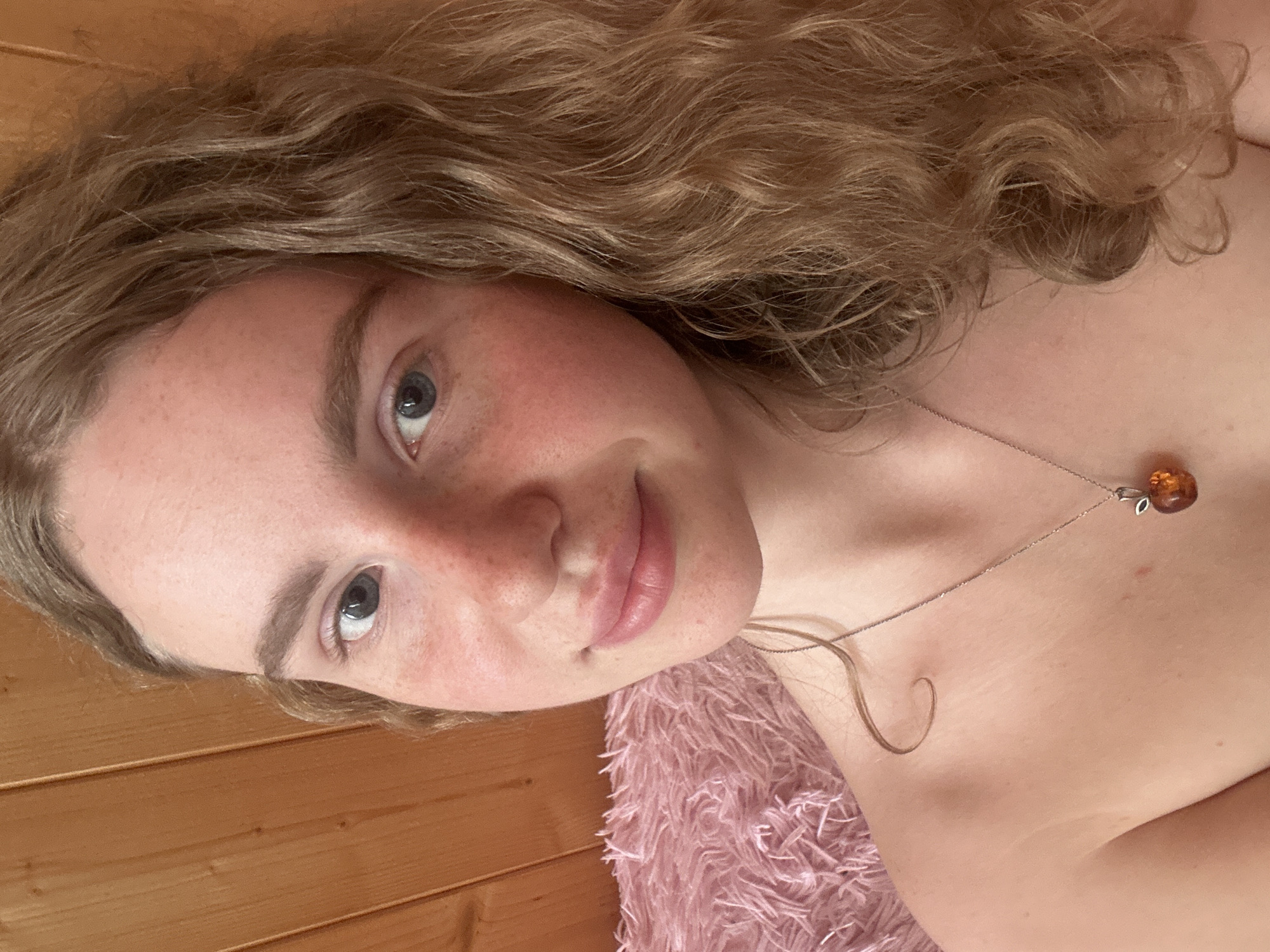 This is a vertically oriented portrait of a young woman with fair skin, light brown to blonde wavy hair, and blue eyes. She has scattered freckles across her nose and cheeks and looks directly at the viewer with a soft, gentle smile. She appears bare-shouldered, wearing a silver-colored chain necklace with a small, amber-toned pendant. Her wavy hair falls around her face and shoulders.
The background on the left side features light brown wooden paneling, while the lower right foreground shows a soft, textured pink material, likely a fluffy blanket or pillow. This suggests a cozy, indoor setting. The woman's expression conveys a sense of calm and serenity. There is no visible text in the image. The location context for this image is Vasilyevo, Russia.