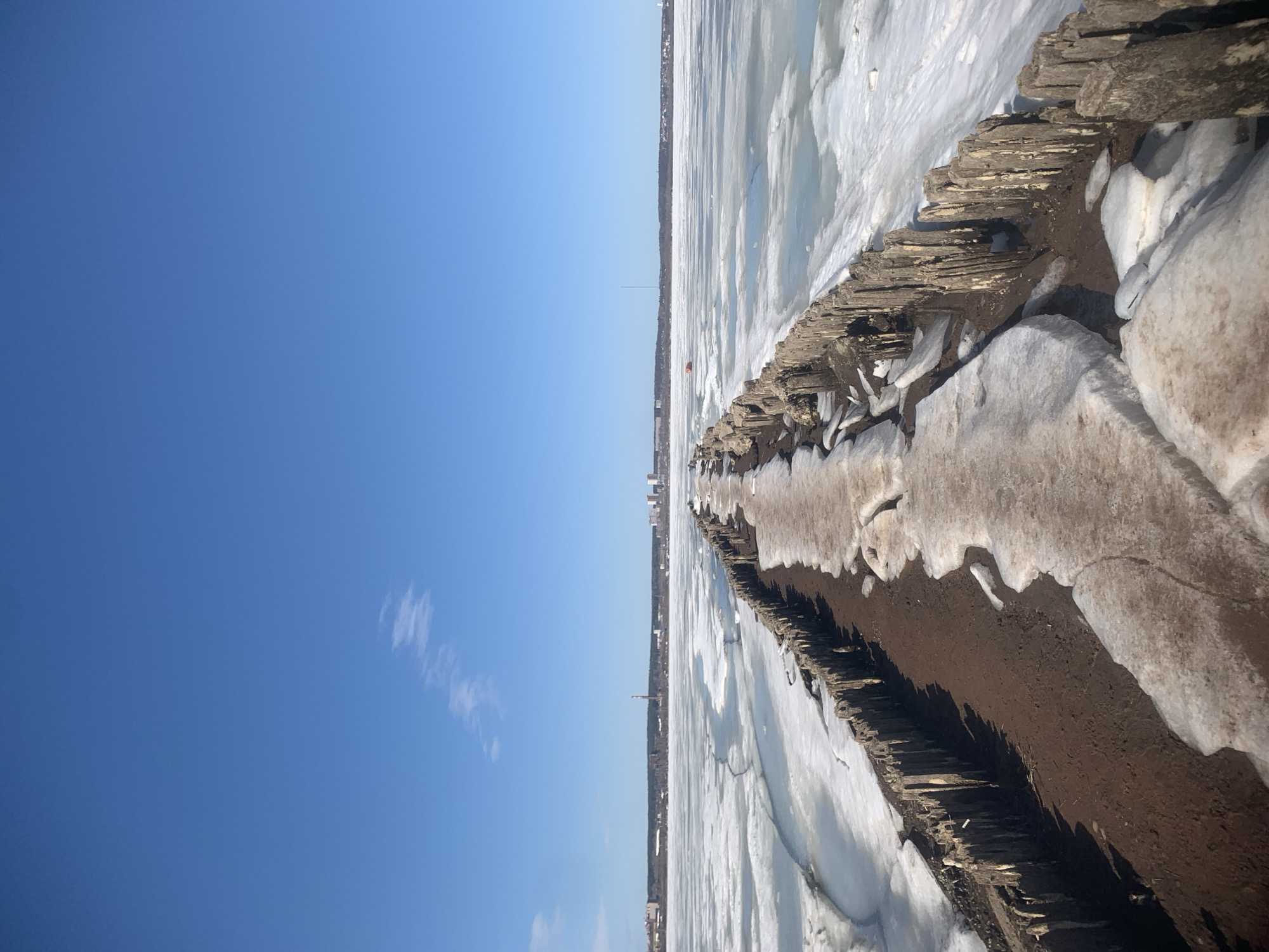 The image shows a pier or breakwater covered in ice and snow, with a dirt path along the center. The structure is made of wood and rocks. The surrounding area is a frozen body of water. In the distance, the skyline of Izhevsk, Russia is visible. A communication tower is noticeable on the horizon. The sky is blue with a few clouds. The image appears to have been taken on a clear day.