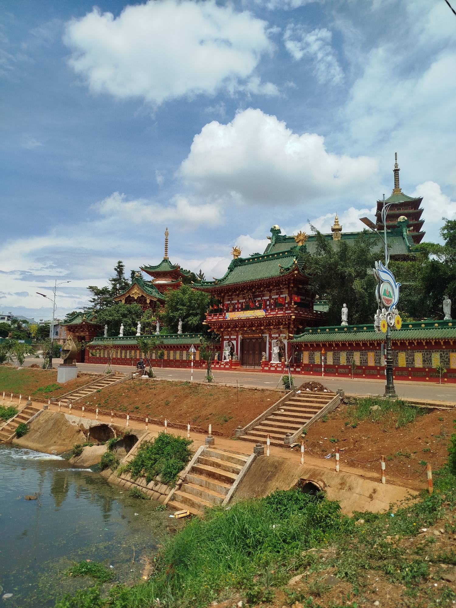 The image depicts a temple complex in Pleiku Ward, Vietnam. The complex is characterized by traditional East Asian architecture, featuring a multi-tiered pagoda, green-tiled roofs with golden embellishments, red walls, and statues along the perimeter. A stream flows in the foreground, with concrete embankments and steps leading up to the temple grounds. The street in front of the temple has a decorative lamppost, and there are trees interspersed throughout the complex. The sky is blue with scattered white clouds. A person can be seen on a scooter in front of the temple. The immediate foreground contains greenery and some visible debris in the water. The ground around the temple is earthen, with some exposed soil.