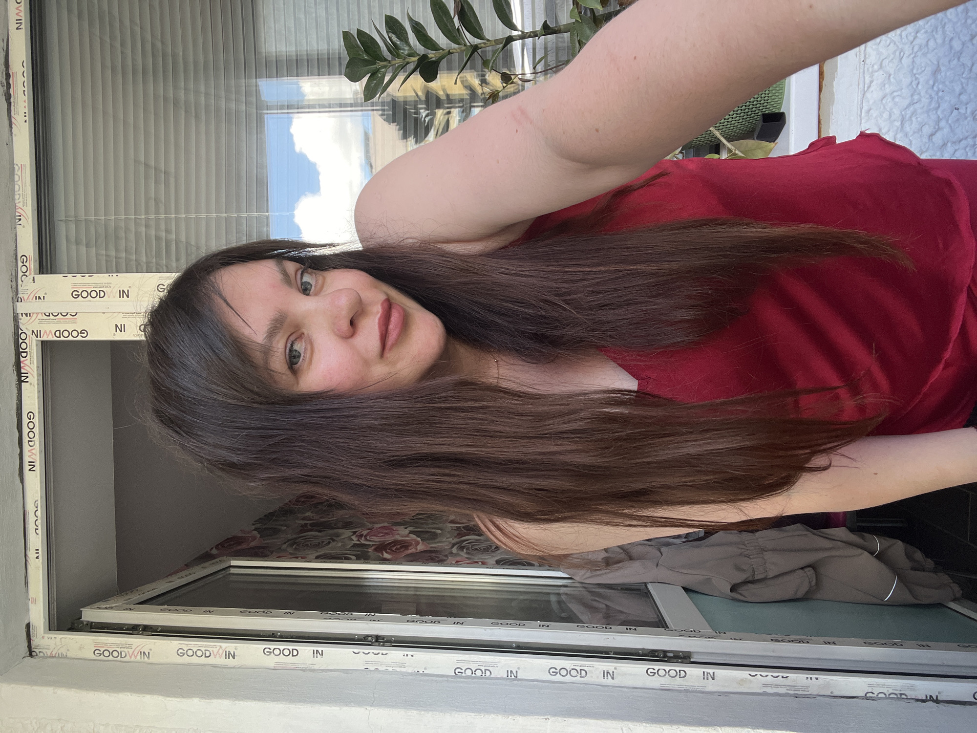 In Noviy, Russia, a young woman with long, dark brown hair and light blue eyes is pictured in a selfie taken outdoors near a window. She is wearing a red top. The background shows a window with blinds, a plant on the windowsill, and part of a doorway with wallpaper that has a floral pattern. The window frame has "GOODWIN" printed on it.