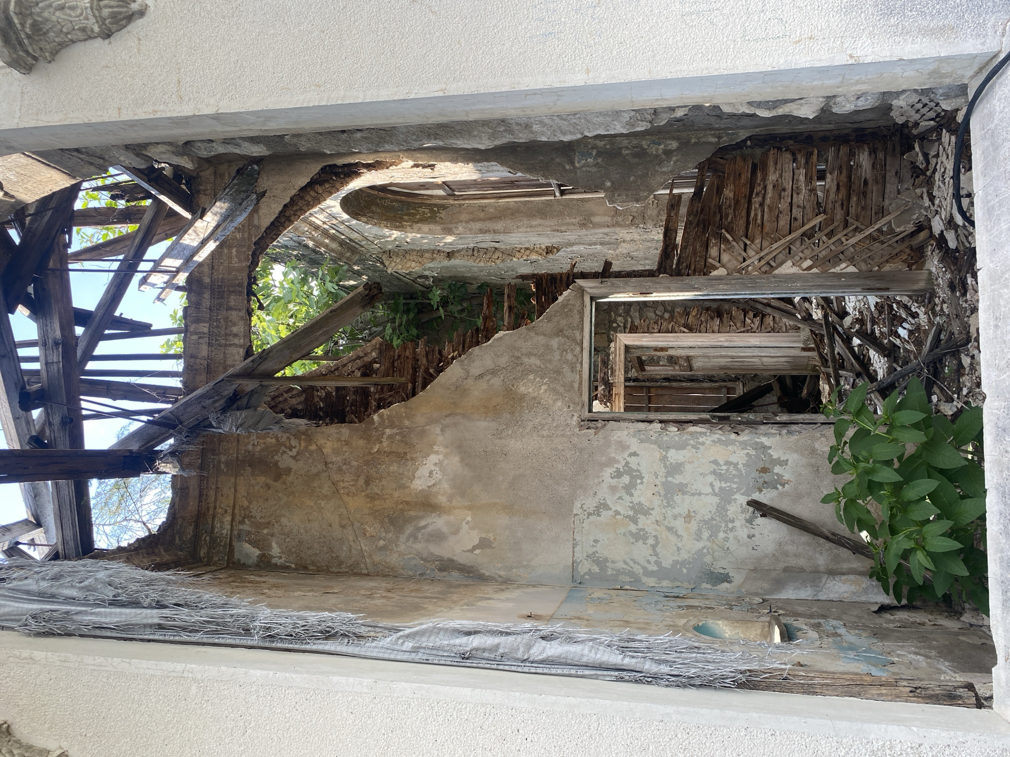 The image depicts the interior of a heavily damaged, possibly abandoned building in Psakhara, Abkhazia. The structure shows signs of significant decay and collapse, with a ruined roof exposing the sky above, supported by a network of broken wooden beams. The walls are crumbling, revealing plaster and brickwork, and some sections are covered in peeling paint. A damaged staircase is visible, constructed from splintered wood and debris. There are multiple doorways, some leading into darker, unknown spaces. Green foliage is growing through the rubble and within the structure, indicating a lack of human activity and the encroachment of nature. The overall impression is one of severe disrepair and neglect.
