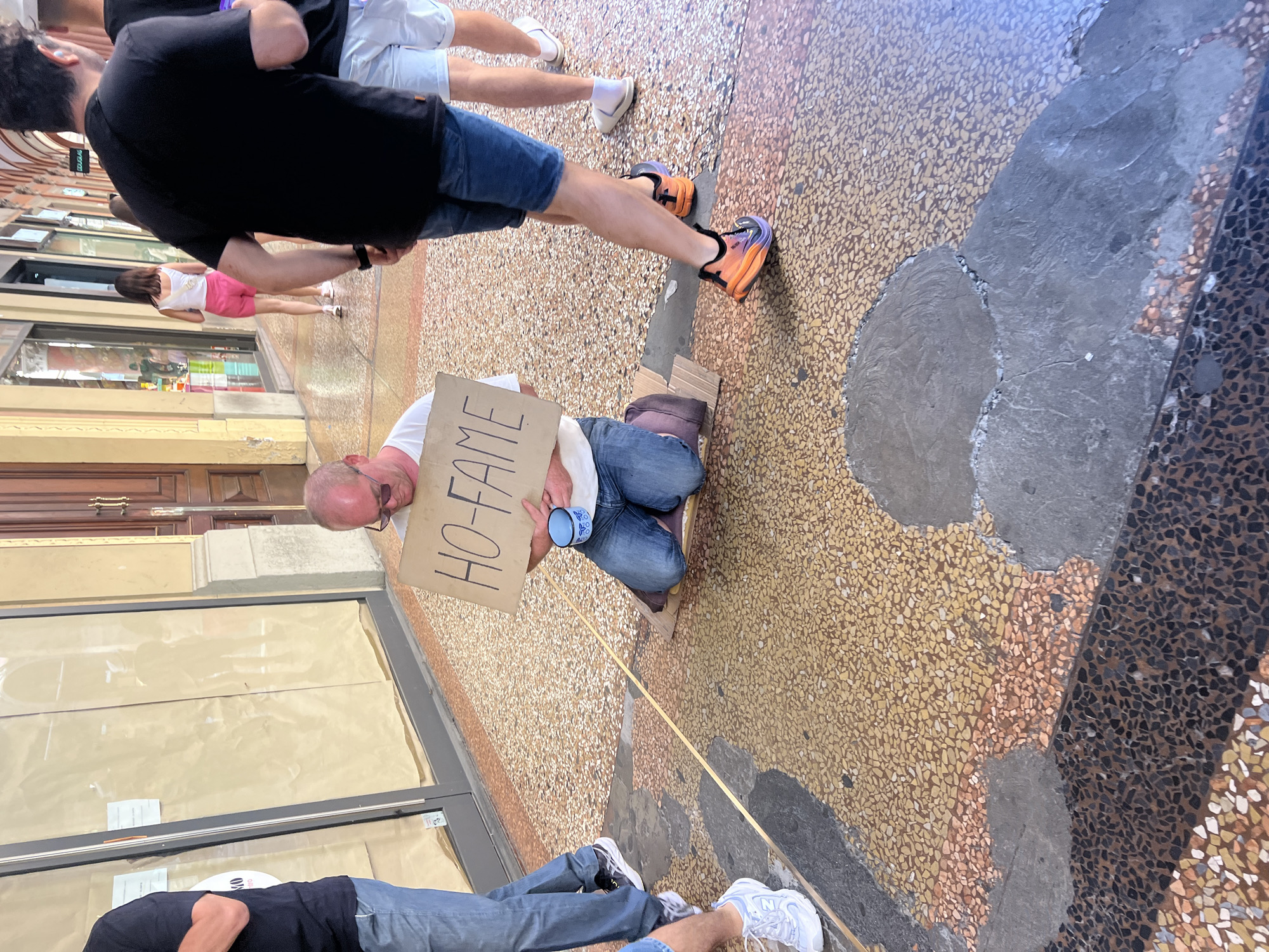 A street scene shows a man sitting on the ground holding a sign that reads "HO-FAME" (I am Hungry). He is wearing a white shirt, blue jeans, and glasses. He is sitting on a piece of cardboard with a blue mug next to him. Several people are walking past him. The street is made of small yellow and brown tiles. The scene is in Unknown, Unknown.