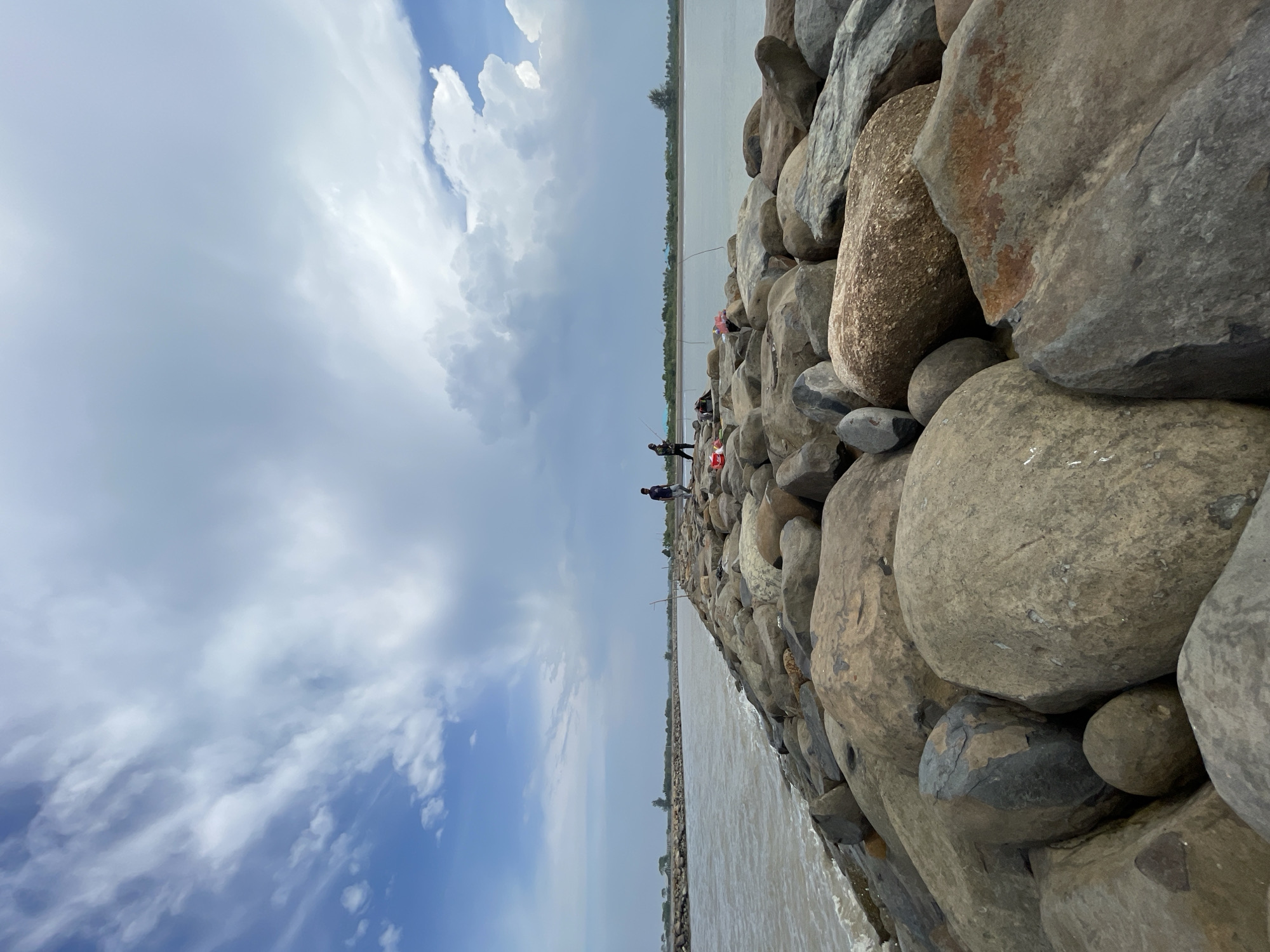 The image shows a rocky jetty extending into a body of water under a cloudy sky. The jetty is constructed of large, irregular stones of various sizes and colors, with a path worn into the top. Several people are visible on the jetty, some appear to be fishing with rods, while others are standing or sitting. One person is walking on the far end of the jetty, and a few individuals are clustered further down. The water is choppy and a light brown color. The background shows a distant shoreline with trees and a few buildings. The sky is filled with dramatic, white and grey clouds, suggesting a dynamic weather system. The overall scene depicts a recreational or working area by the water, likely a breakwater or pier, situated in the vicinity of Kualacangkoi, Indonesia, a region known for its coastal landscapes and fishing activities.