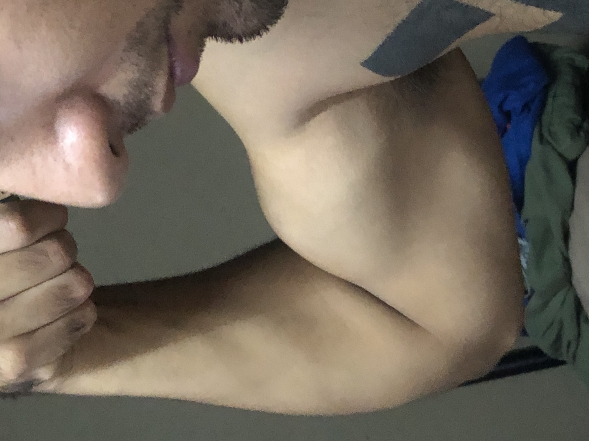 A close-up shot presents a person's face from the nose down to the jawline, along with a flexed right arm exhibiting a prominent bicep. The individual has a stubble beard. The arm appears to have a tattoo near the shoulder. In the background, partially visible, are some items of blue and green clothing. The environment appears to be indoors, possibly a private residence. There is no indication of a location in Buenos Aires, Argentina.